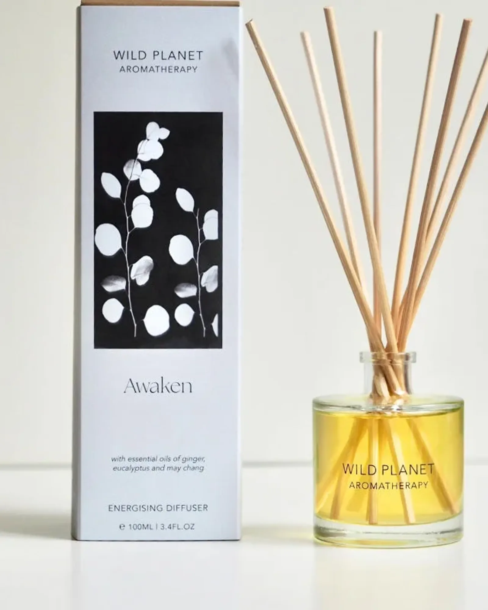 A clear glass bottle of Awaken reed diffuser oil with a fan of natural reeds standing in it, beside its card box printed with the scent's name.