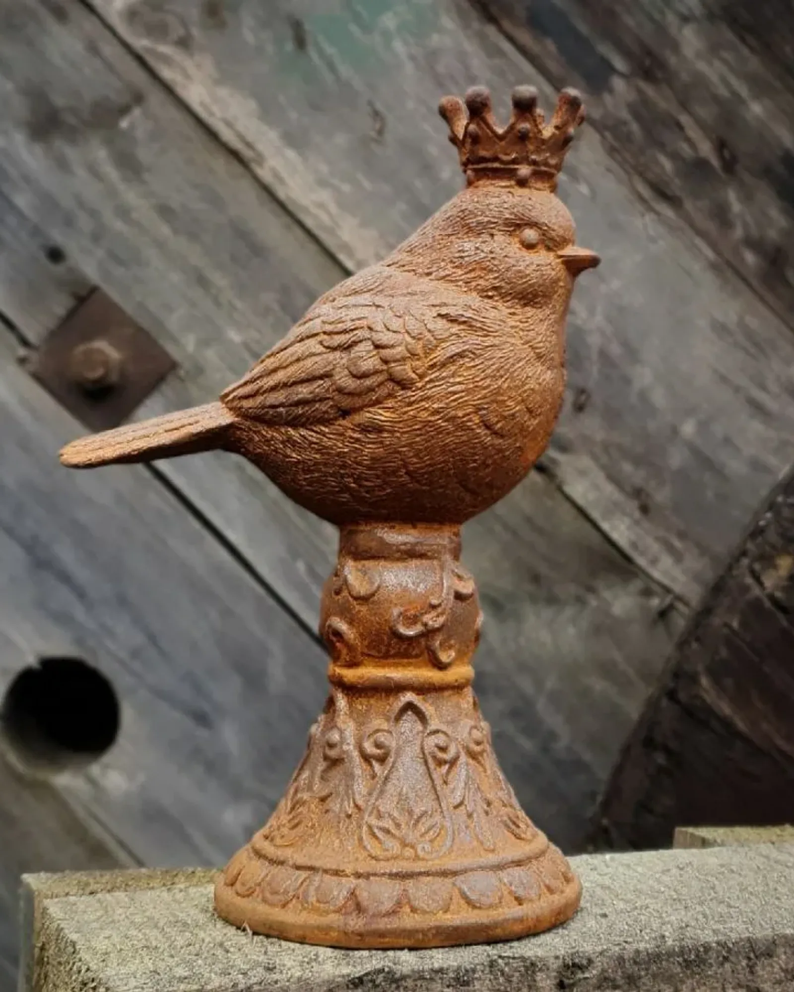 A rusted cast iron songbird wearing a small crown, perched on an ornate bell-shaped pedestal on a fence post.
