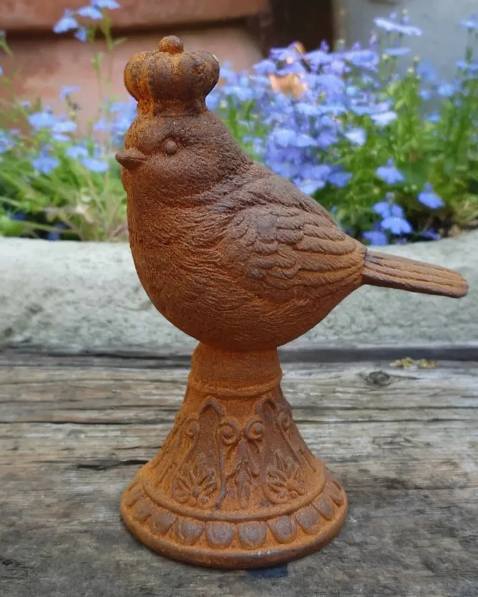 A rusted cast iron bird wearing a crown, perched on an ornate bell-shaped plinth, with blue lobelia behind.