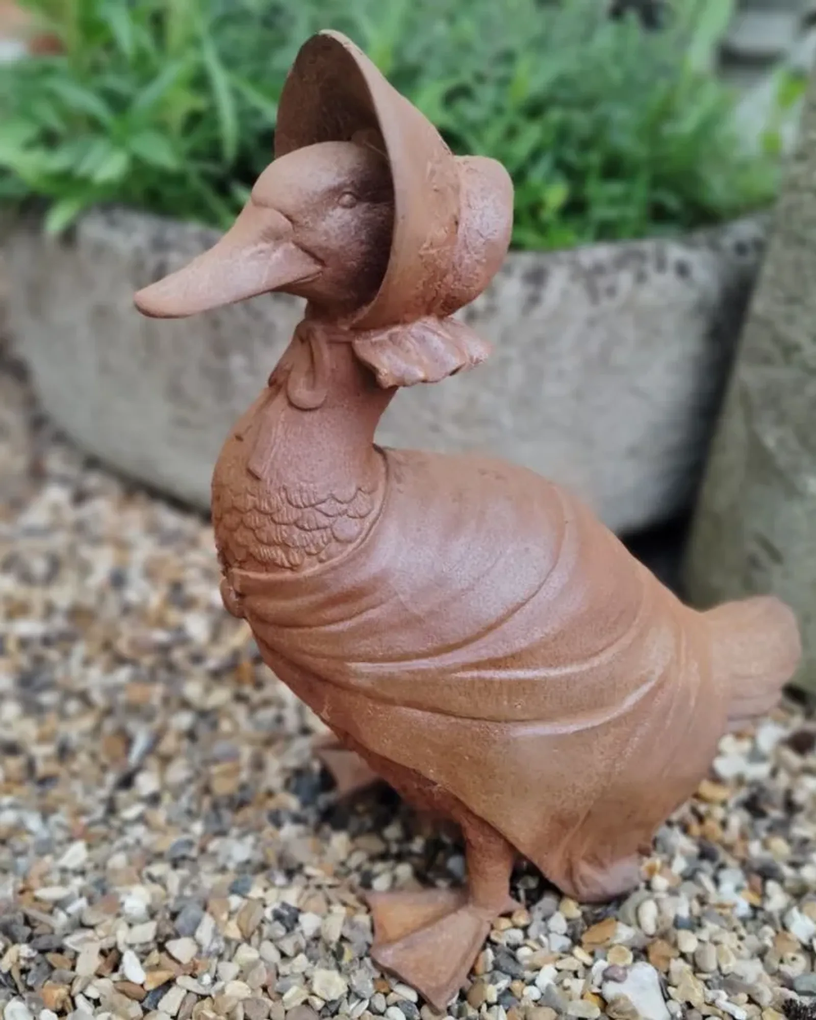 A rusted cast iron duck wearing a bonnet and a shawl, standing on gravel beside a planter.