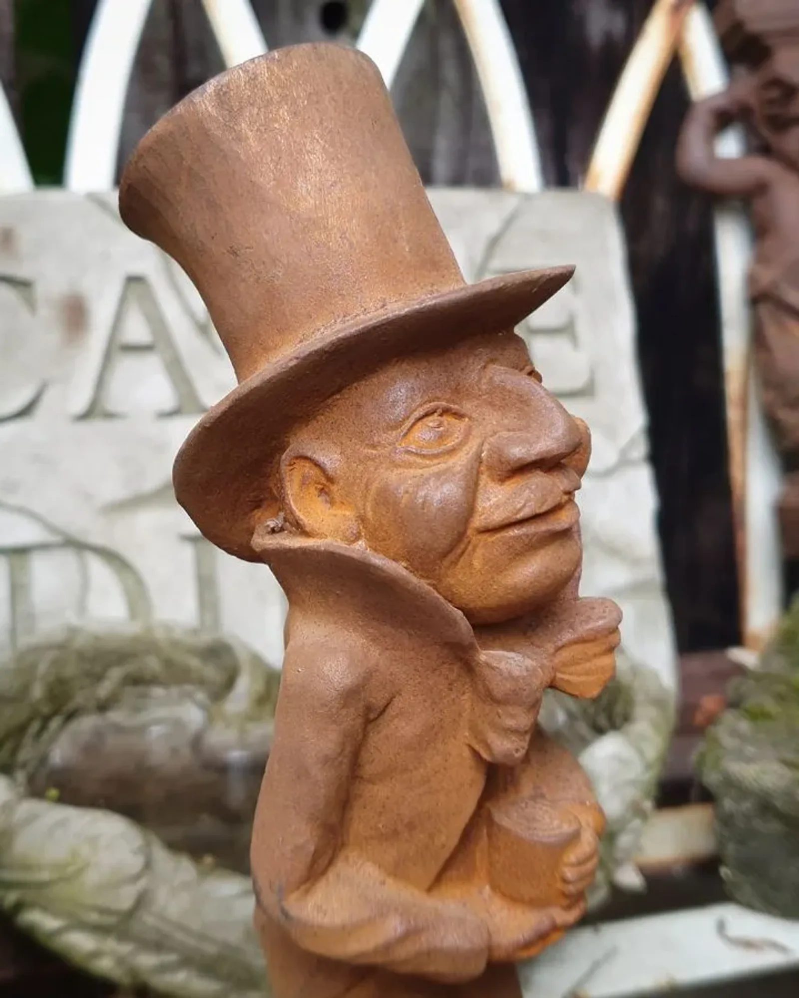 A rusted cast iron figure in a tall top hat and bow tie holding up a cup, photographed among mossy garden ornaments.