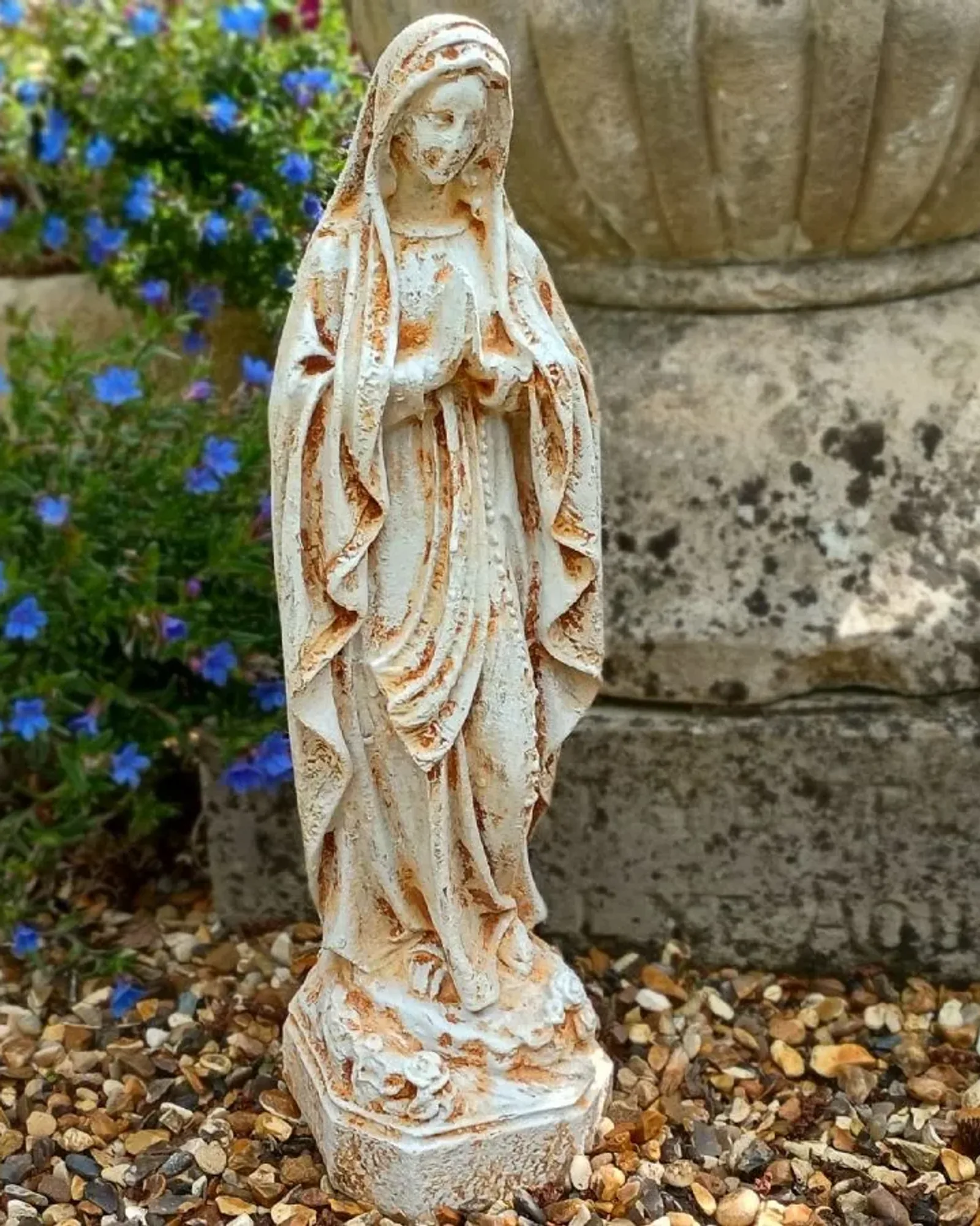 A cast iron statue of Mary with her hands together in prayer, in a distressed white finish with rust showing through, standing on gravel.