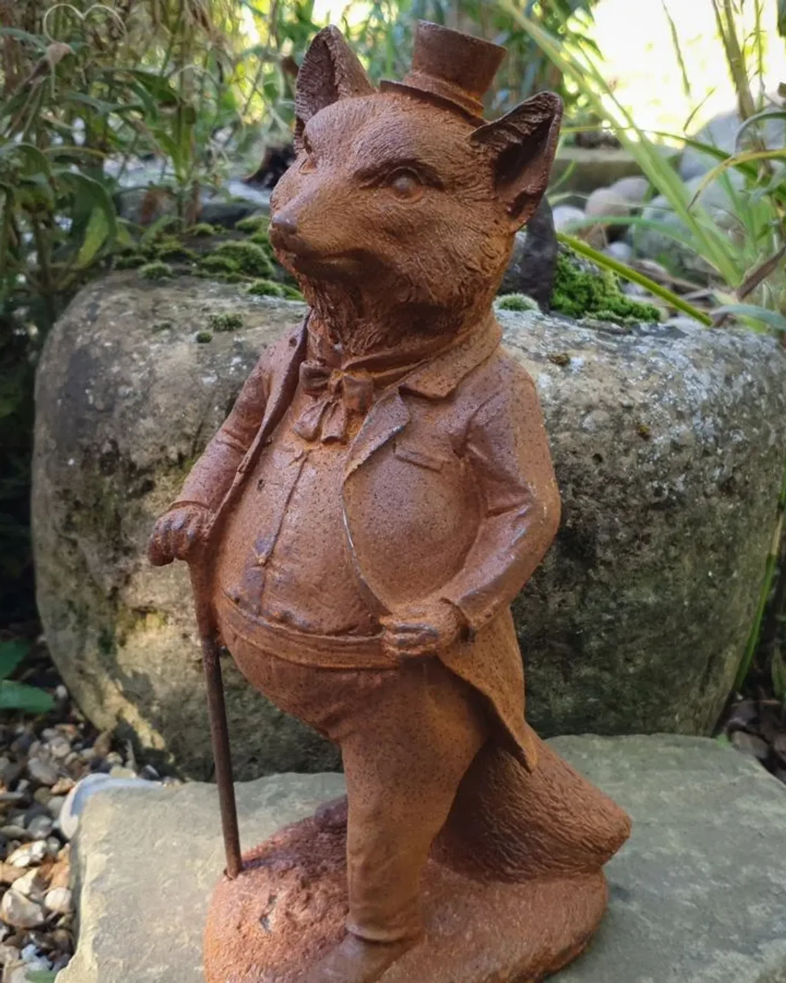 A rusted cast iron fox standing upright in a top hat, jacket and bow tie, leaning on a cane, on an oval base in a garden.