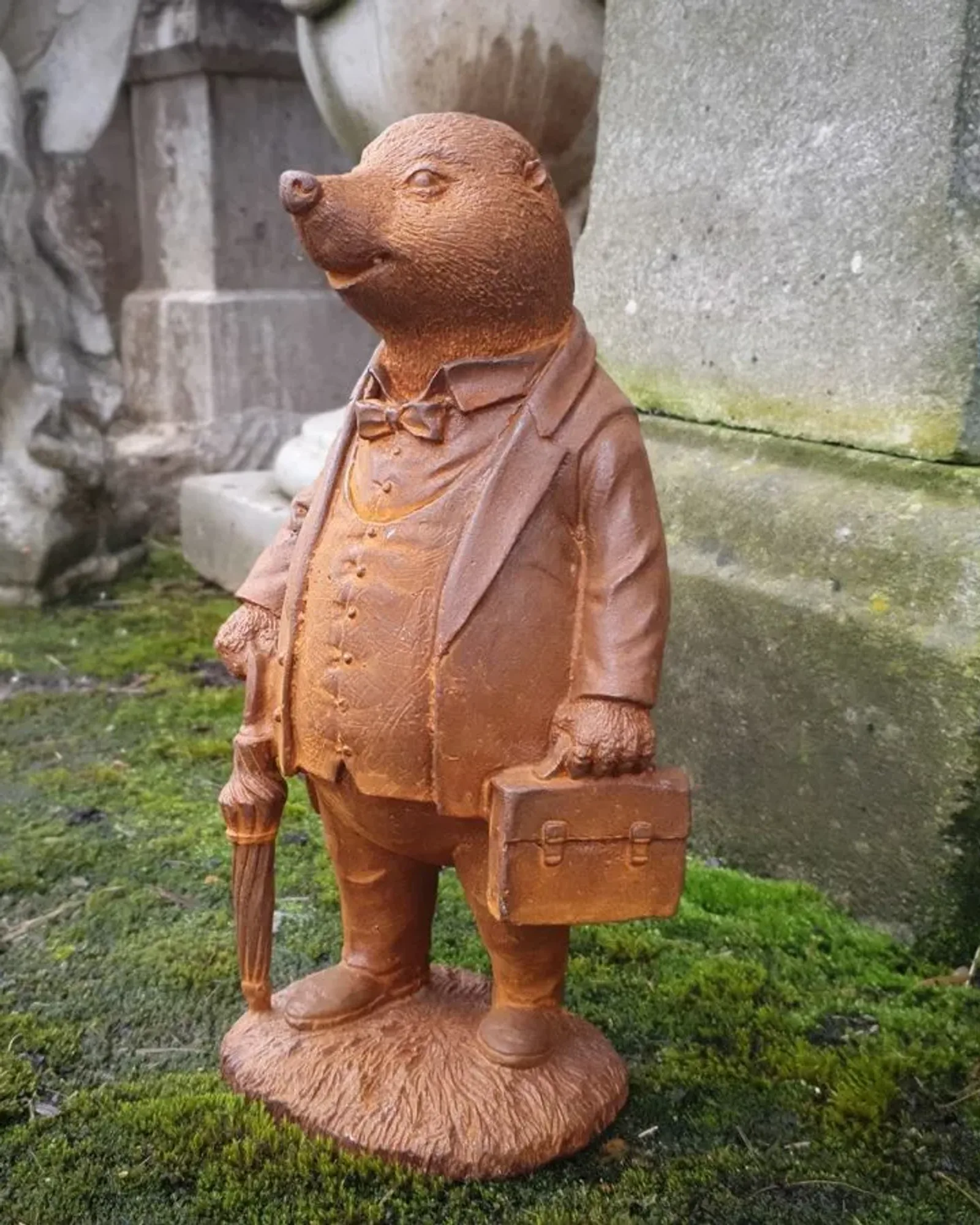 A rusted cast iron mole standing upright in a suit and bow tie, carrying a briefcase and an umbrella, against mossy stone.