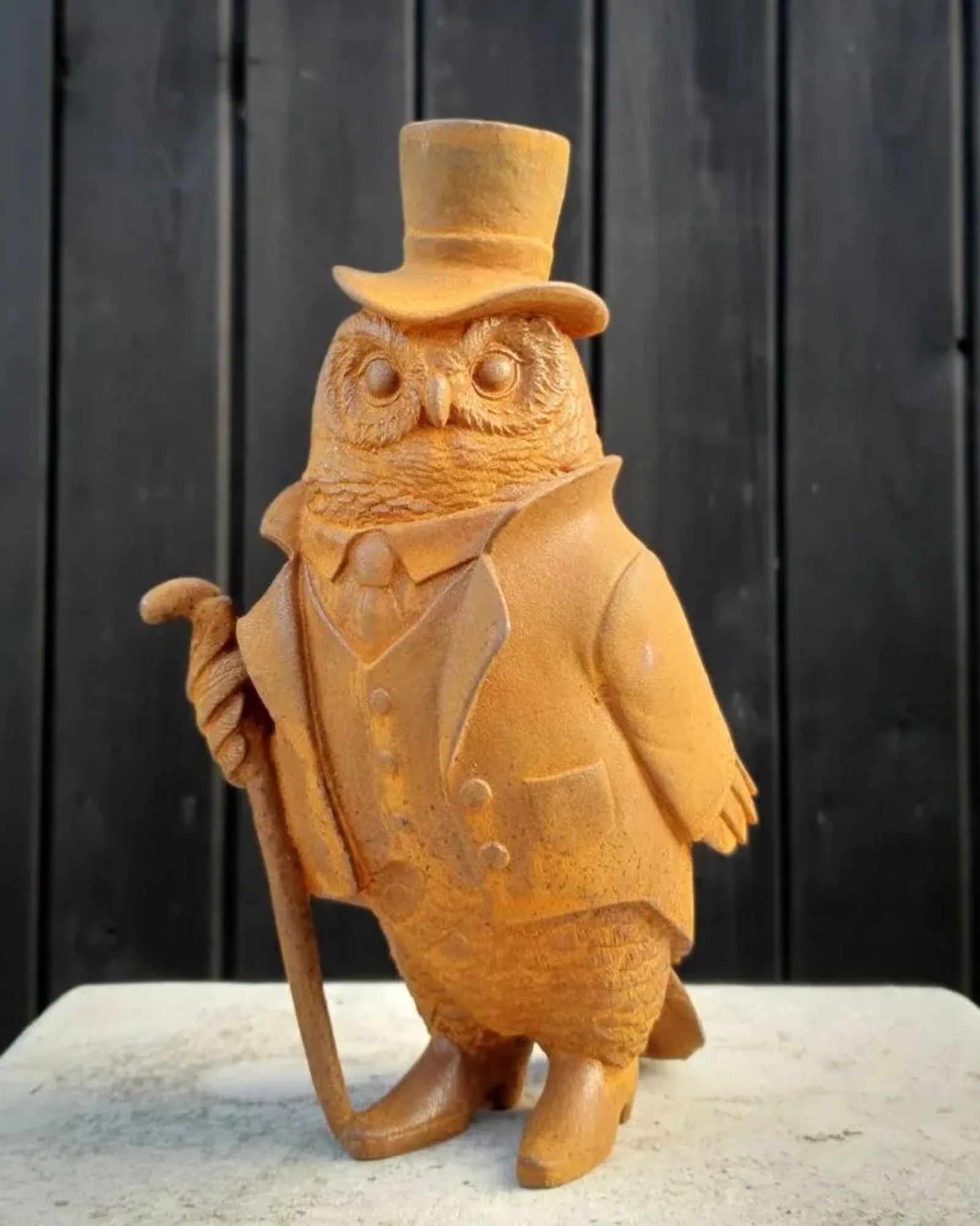 A rusted cast iron owl in a top hat, tailcoat and waistcoat, leaning on a cane, standing on a stone plinth against dark timber.