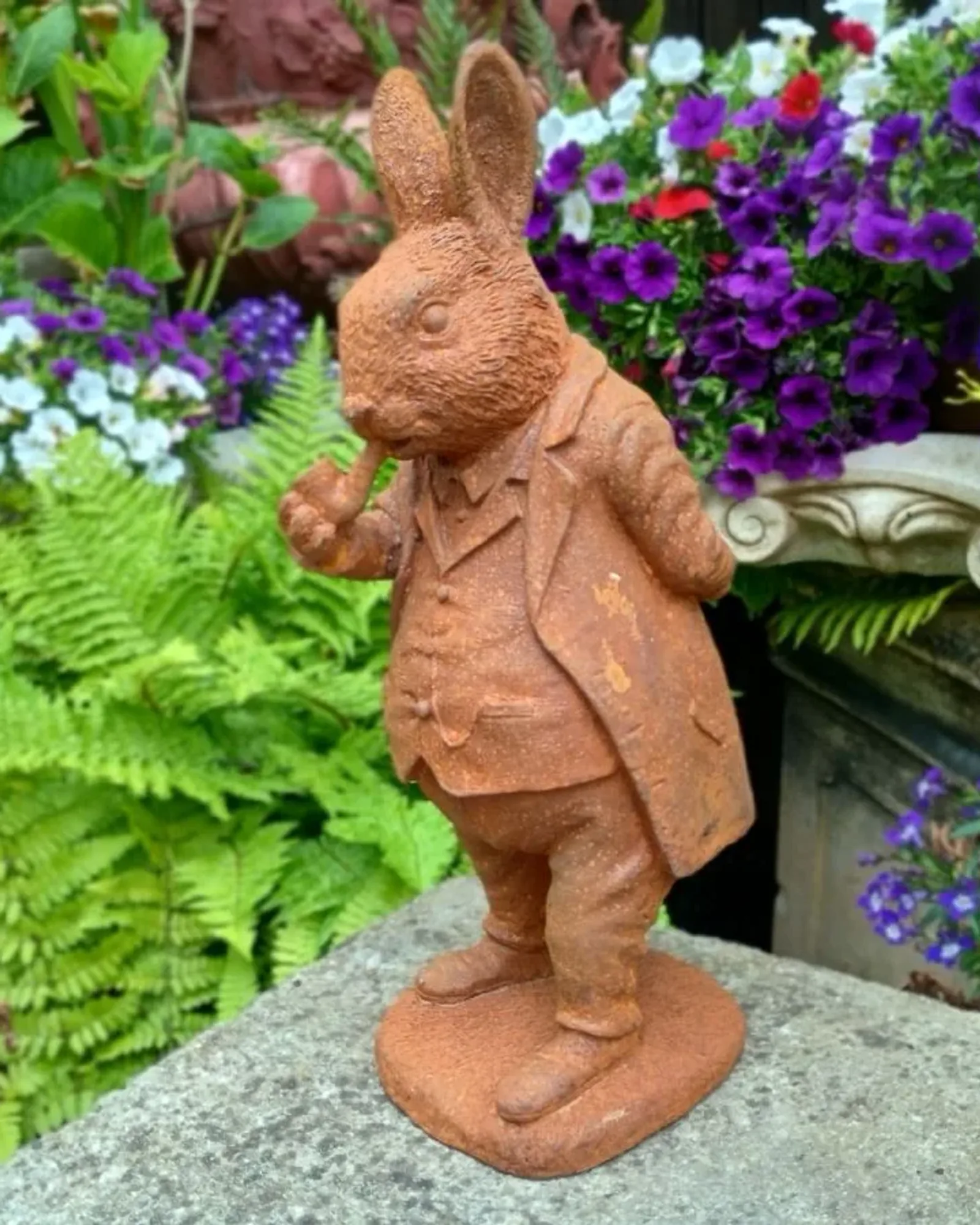 A rusted cast iron rabbit standing upright in a long coat and waistcoat, holding a pipe, on a stone ledge with flowers behind.
