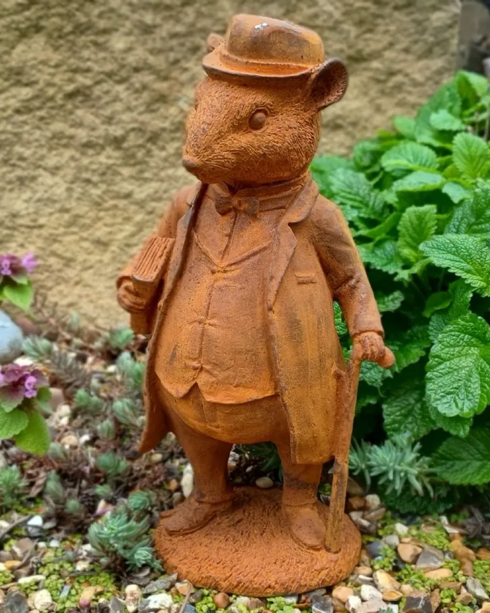 A rusted cast iron rat standing upright in a bowler hat, coat and bow tie, holding a cane with a book under one arm, on a round base among plants and gravel.