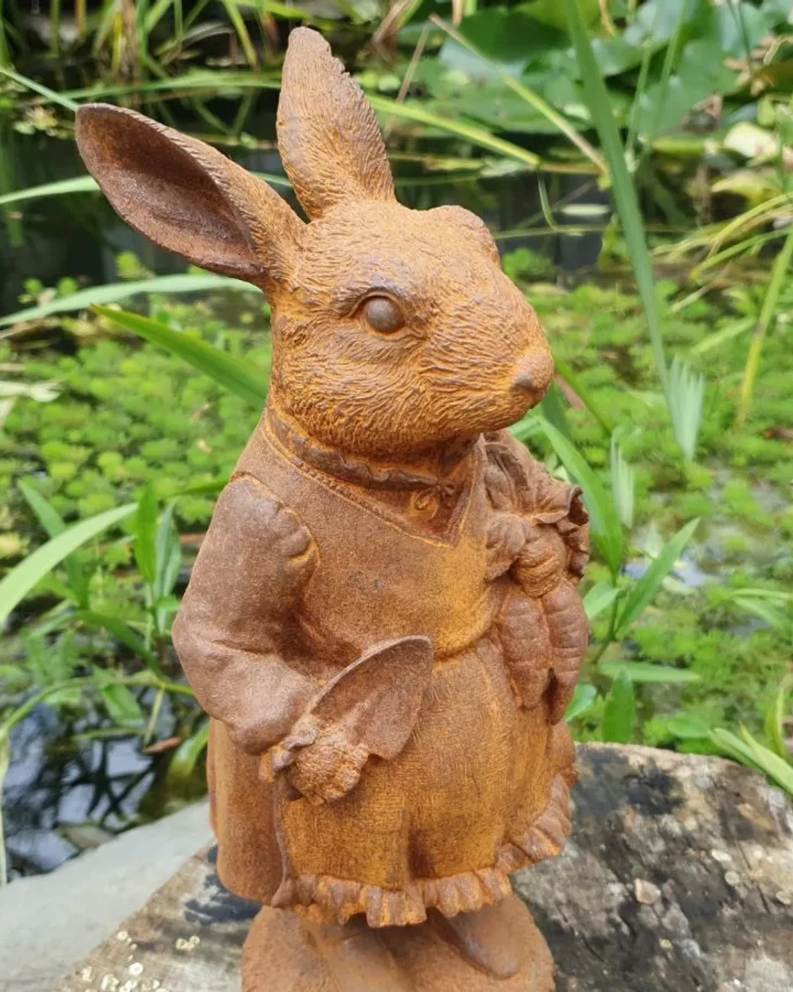 A rusted cast iron rabbit in a frilled dress holding a bunch of carrots, standing on a stump beside a garden pond.