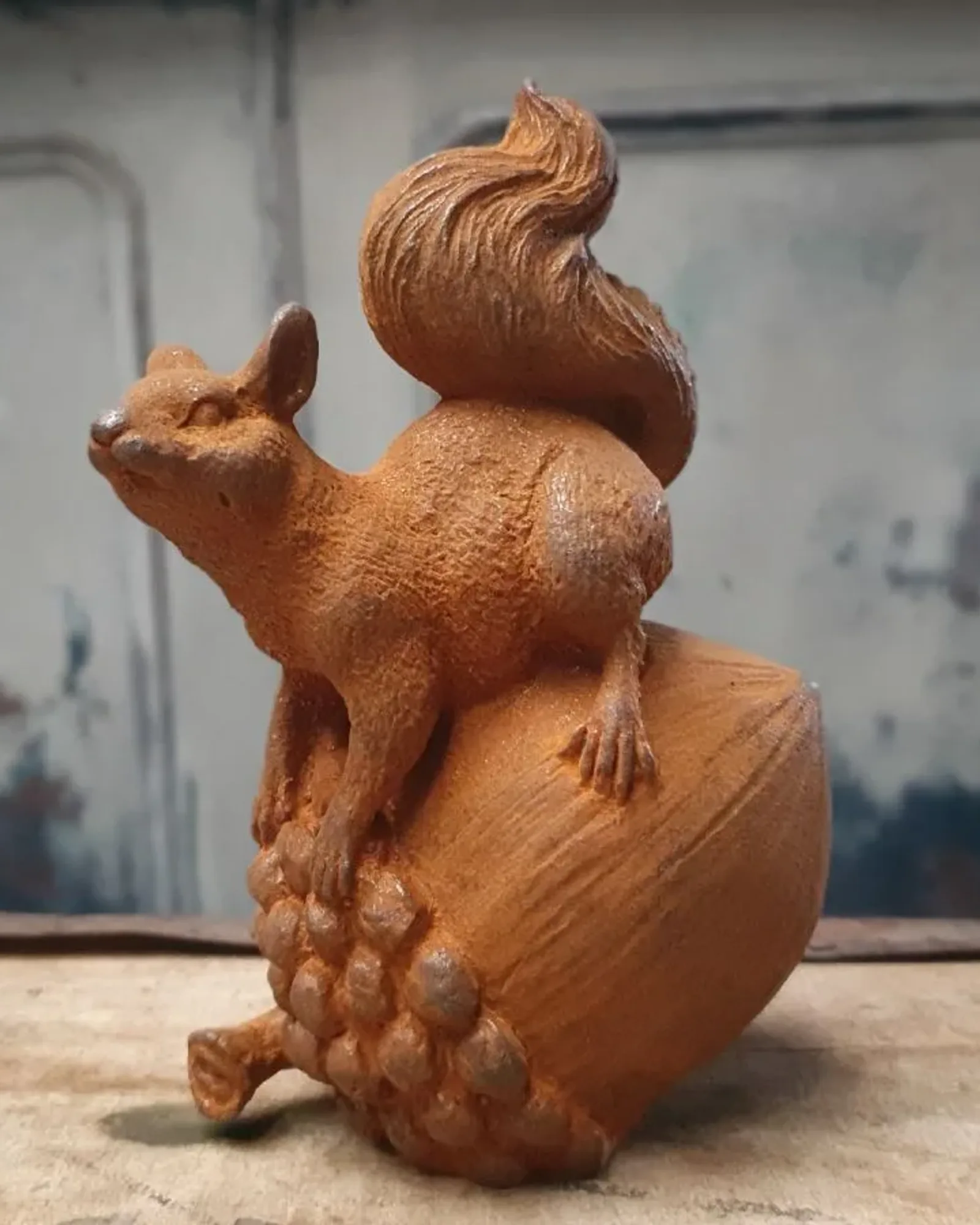 A small rusted cast iron squirrel perched on a nut, photographed against a dark textured wall.