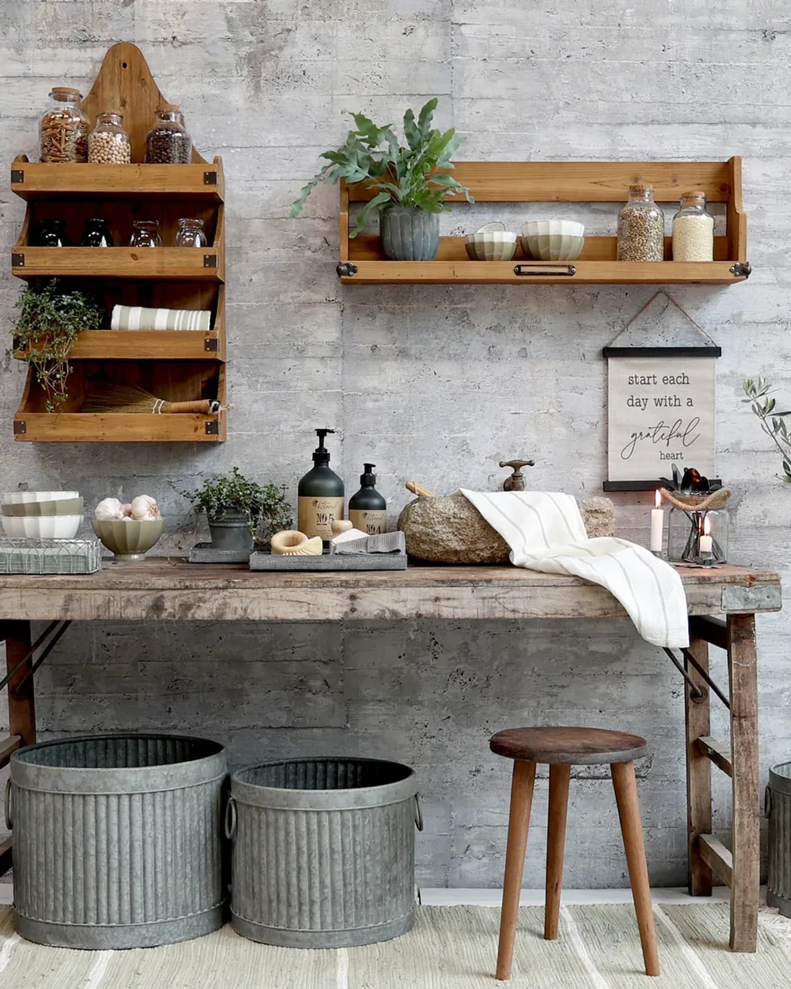 A long natural recycled-wood table with iron corner brackets, loaded with jars, bottles and zinc buckets in a room.