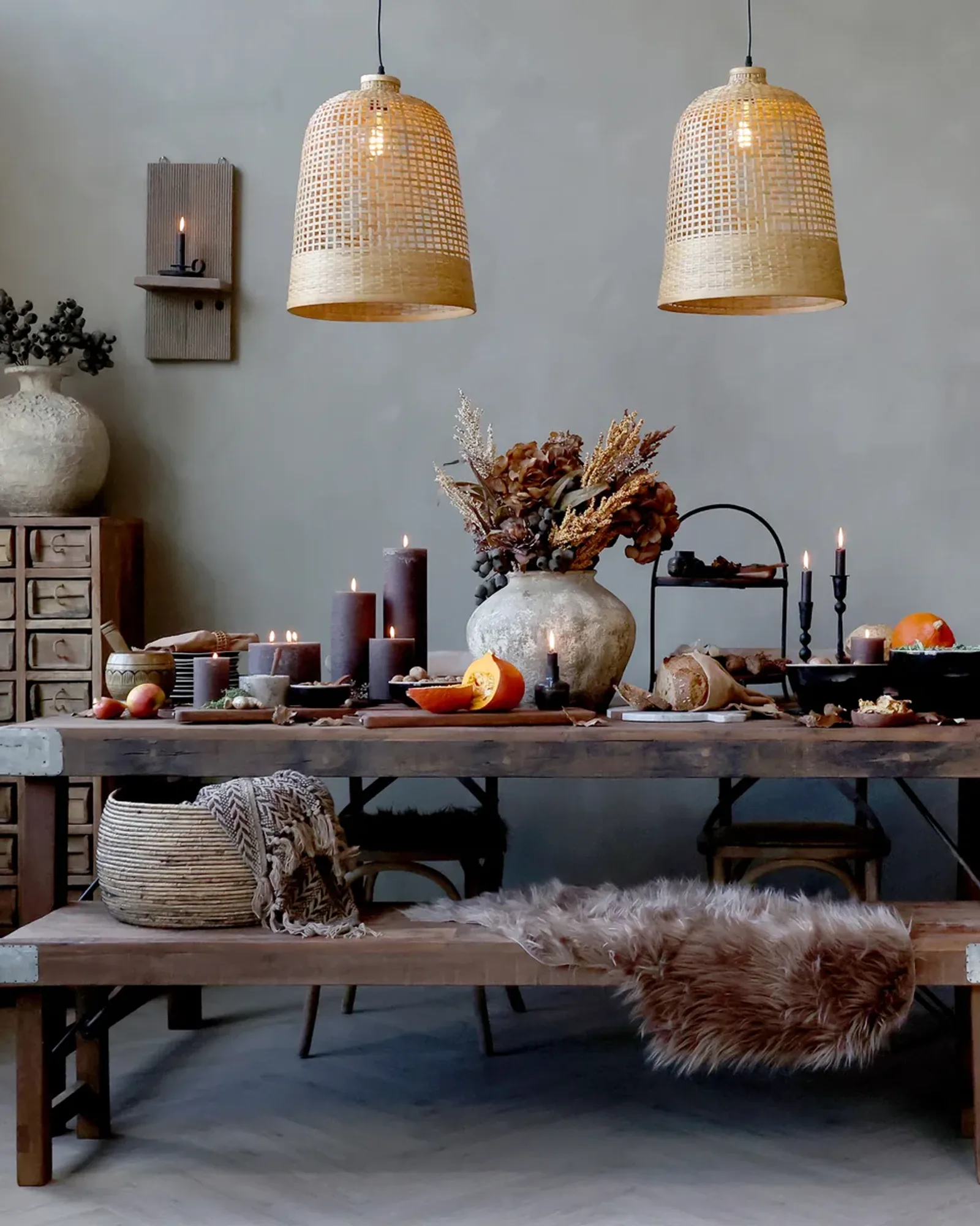 A long reclaimed-wood dining table with iron corner plates and folding cross-braced legs, laid with candles and a vase, a matching bench beside it.