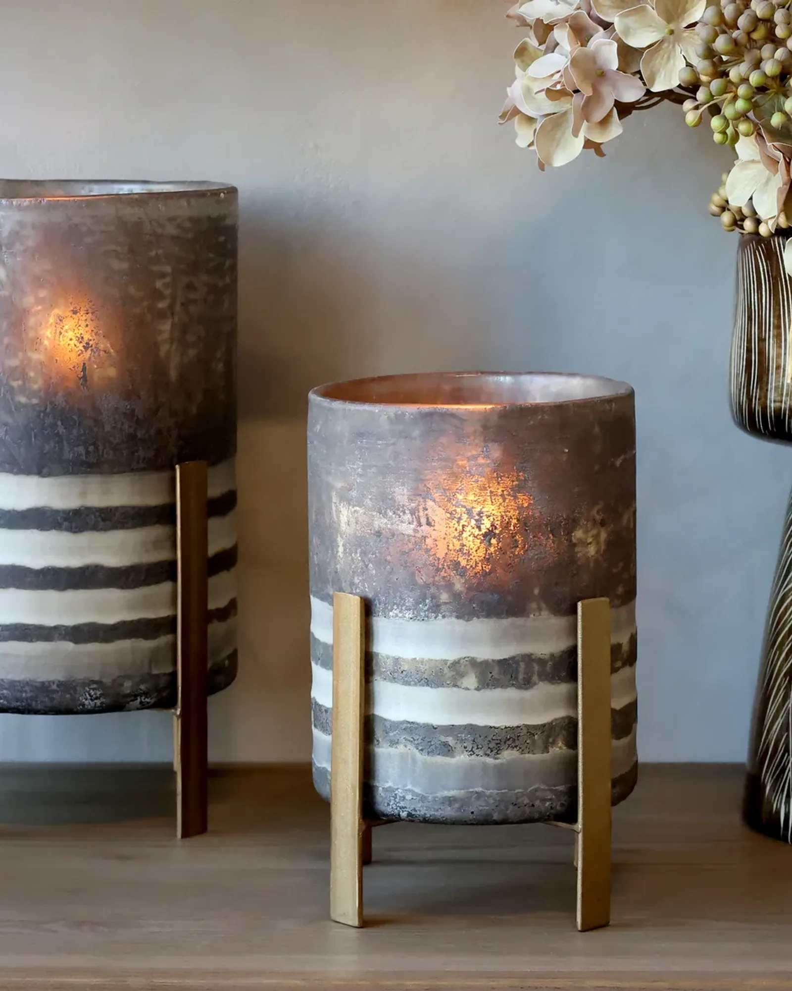 Two coffee-brown glass hurricanes with striped lower bands on brass-coloured metal legs, the smaller at the right beside dried hydrangea.