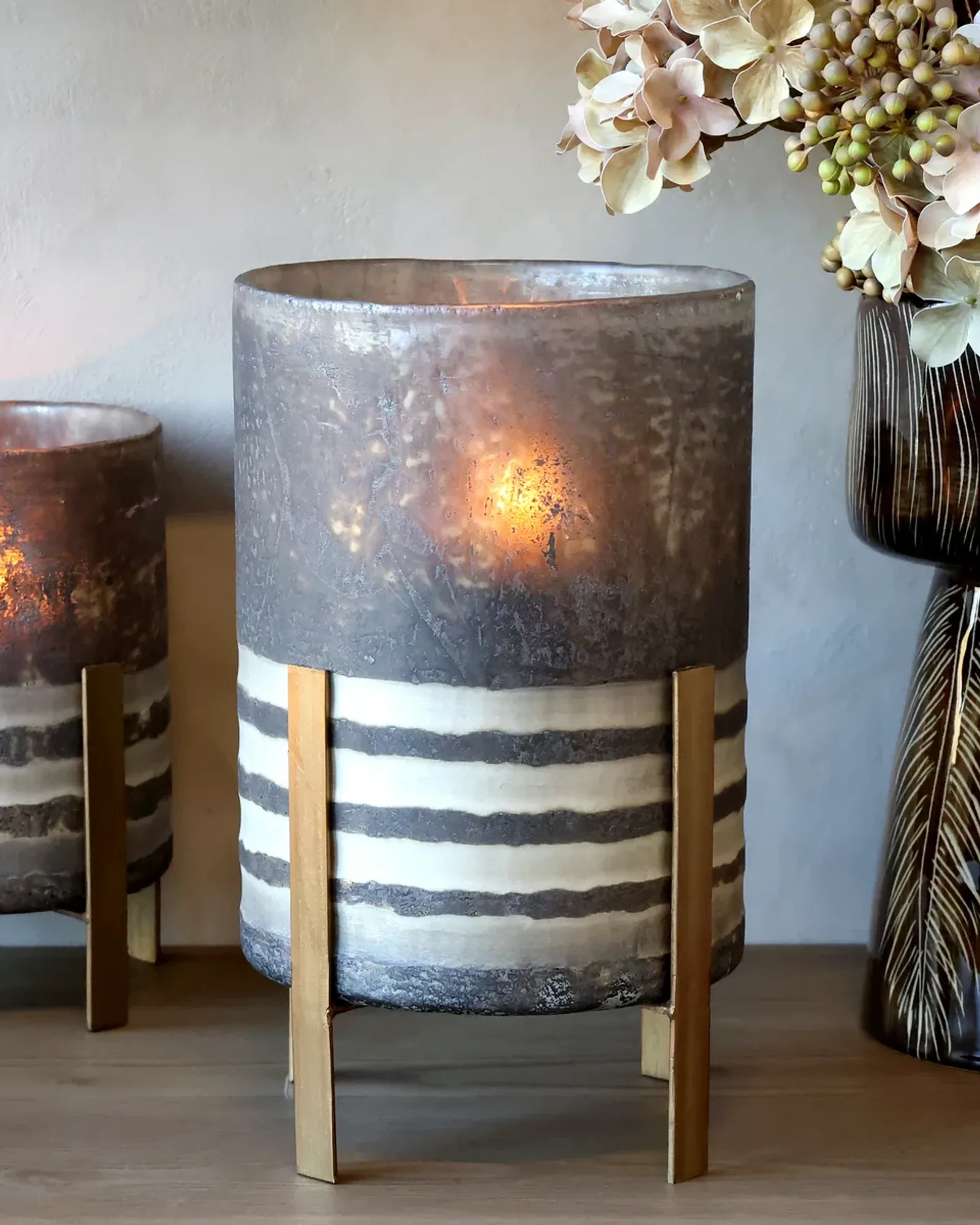 A tall glass hurricane on four brass metal legs, its top antiqued coffee-brown and its lower half banded brown and cream, a candle lit inside and a smaller one at the left.