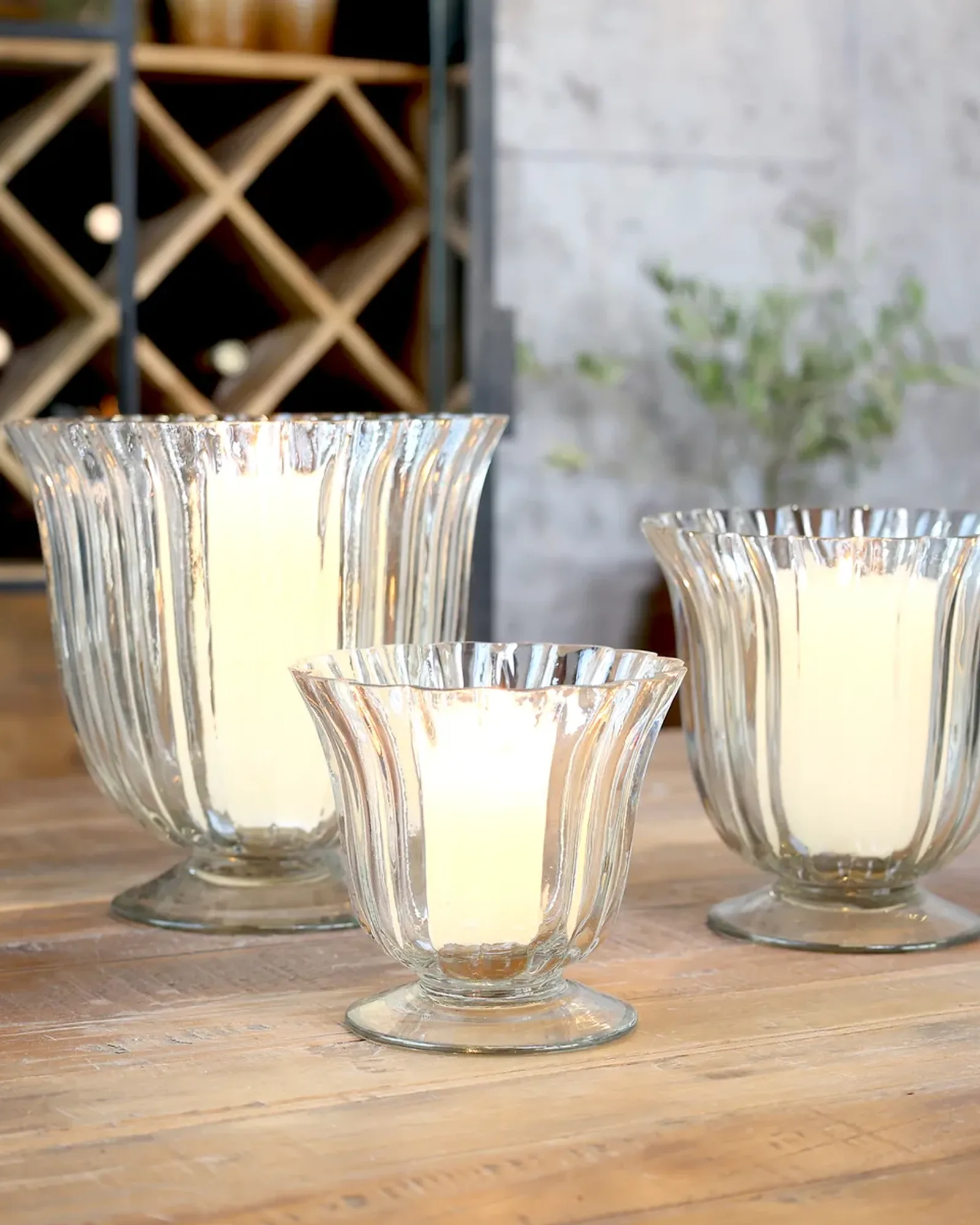 Three clear fluted glass hurricanes on a foot in three sizes on a wooden table, the largest among them.