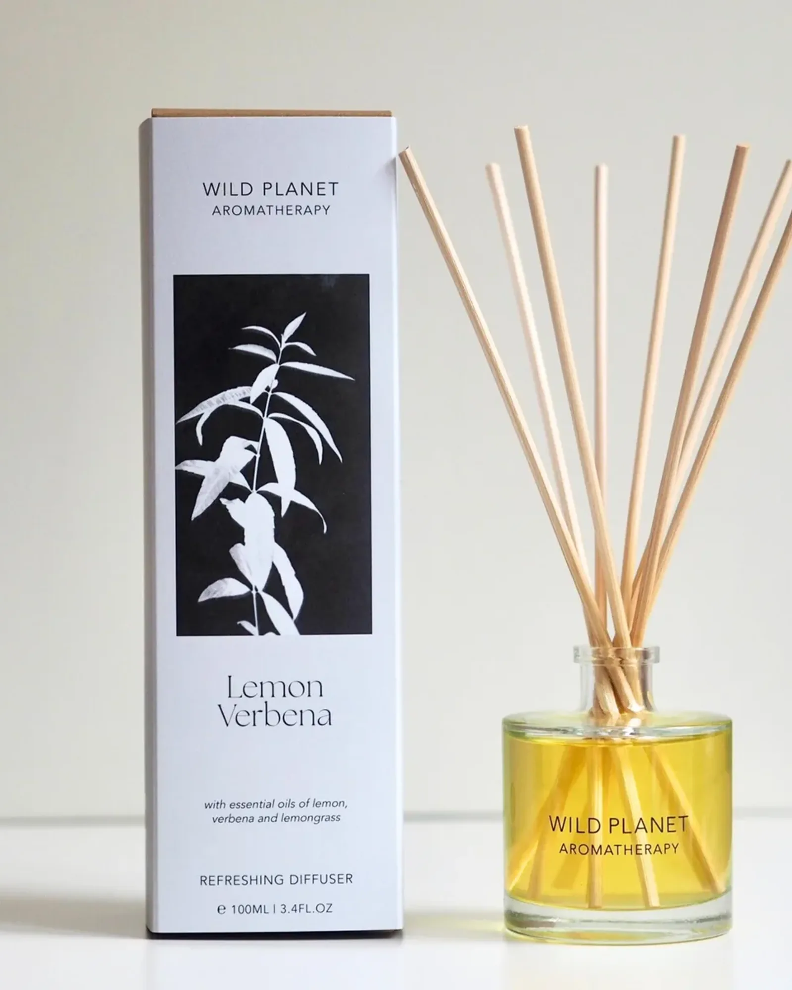 A clear glass bottle of Lemon Verbena reed diffuser oil with a fan of natural reeds standing in it, beside its card box printed with the scent's name.