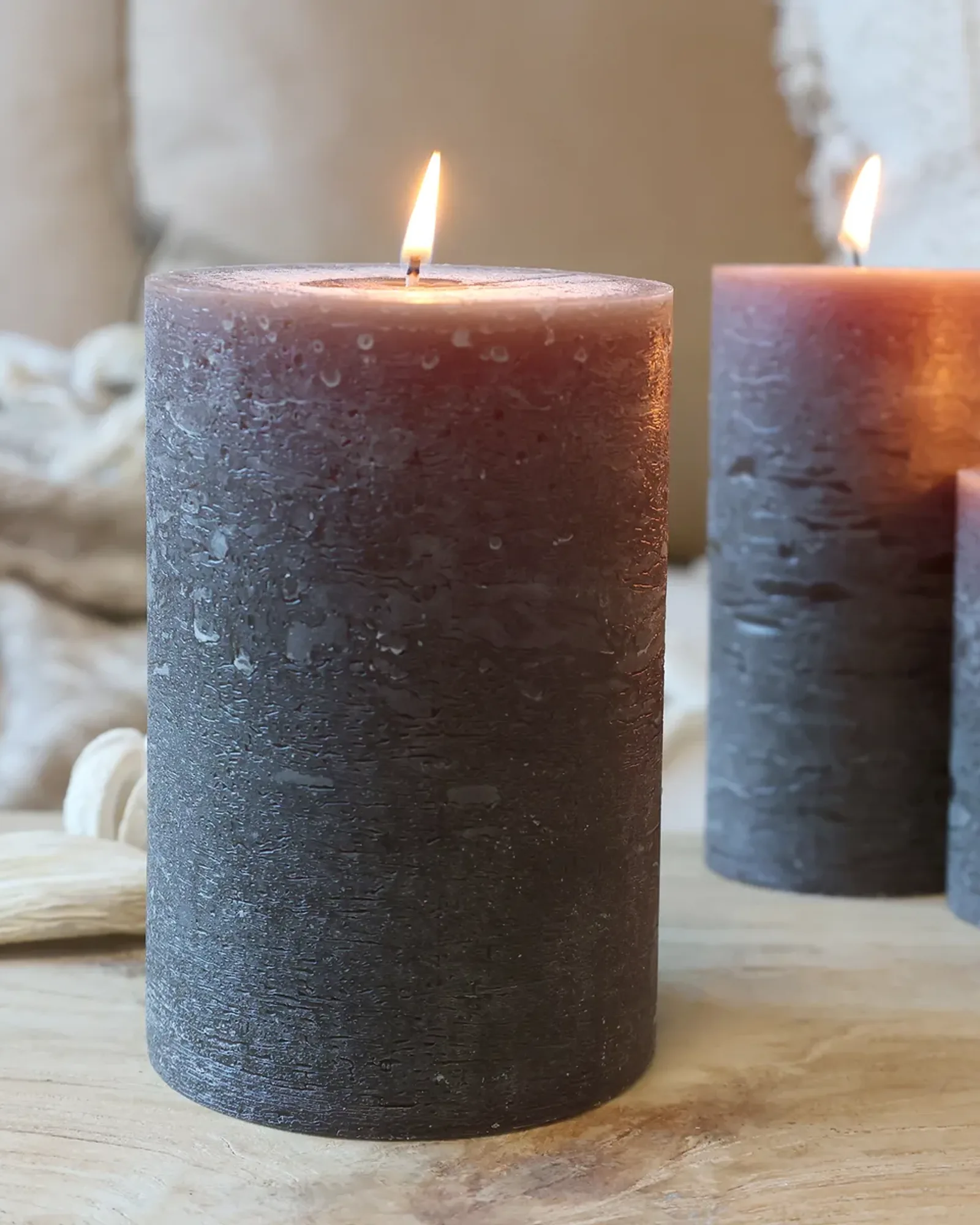 A wide rustic pillar candle in coffee brown, lit, with smaller candles of the same colour behind it.