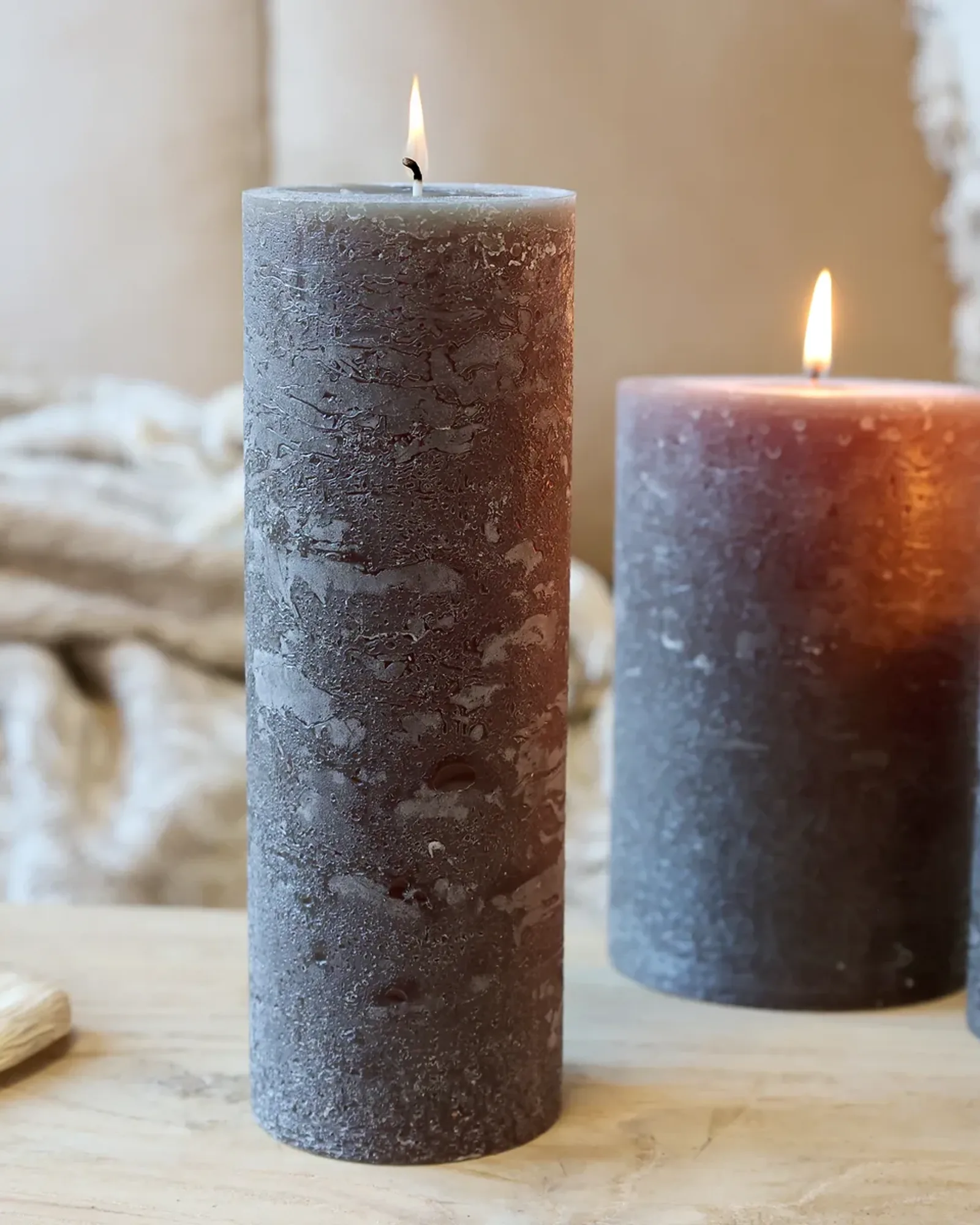A tall, slim rustic pillar candle in coffee brown, lit, with shorter candles of the same colour behind it.