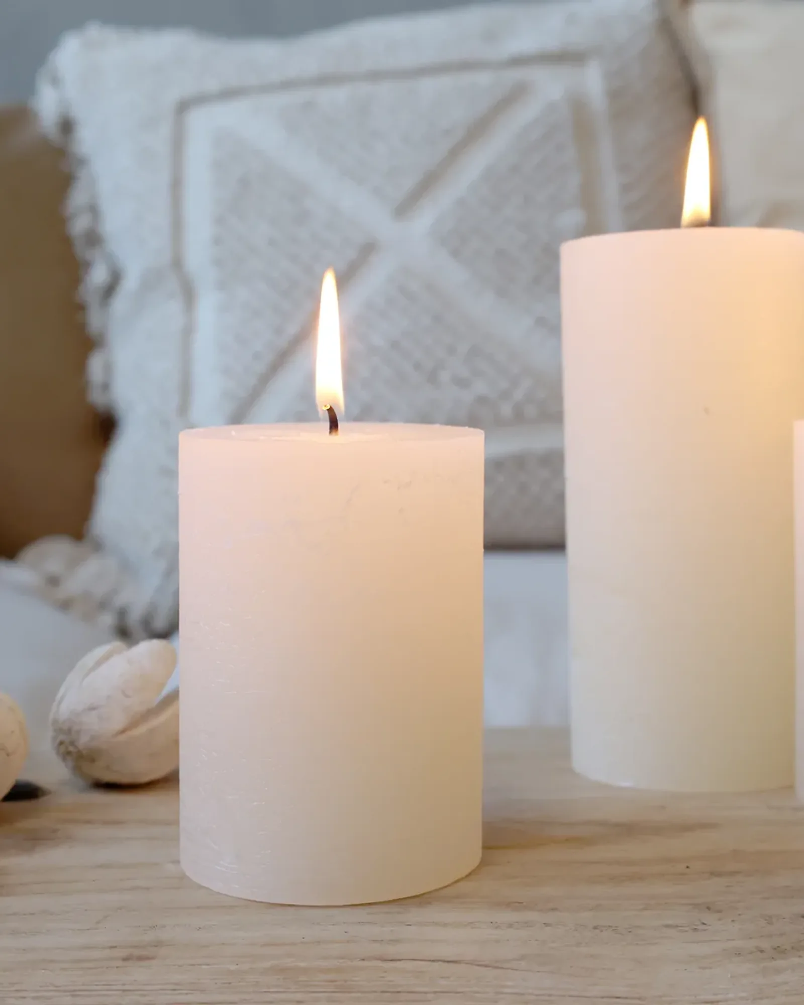 A short rustic pillar candle in cream, lit, with taller candles of the same colour behind it.