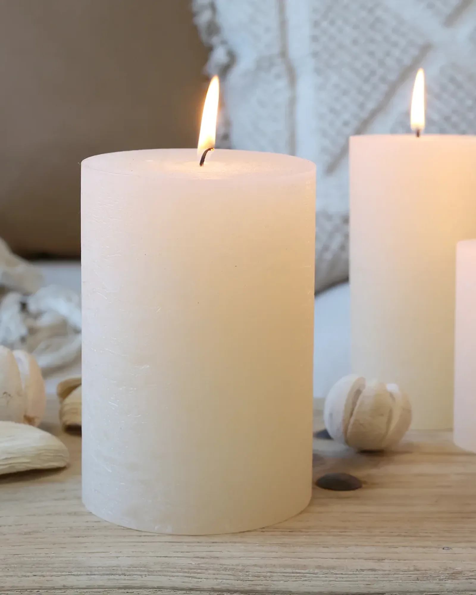 A wide rustic pillar candle in cream, lit, with smaller candles of the same colour behind it.