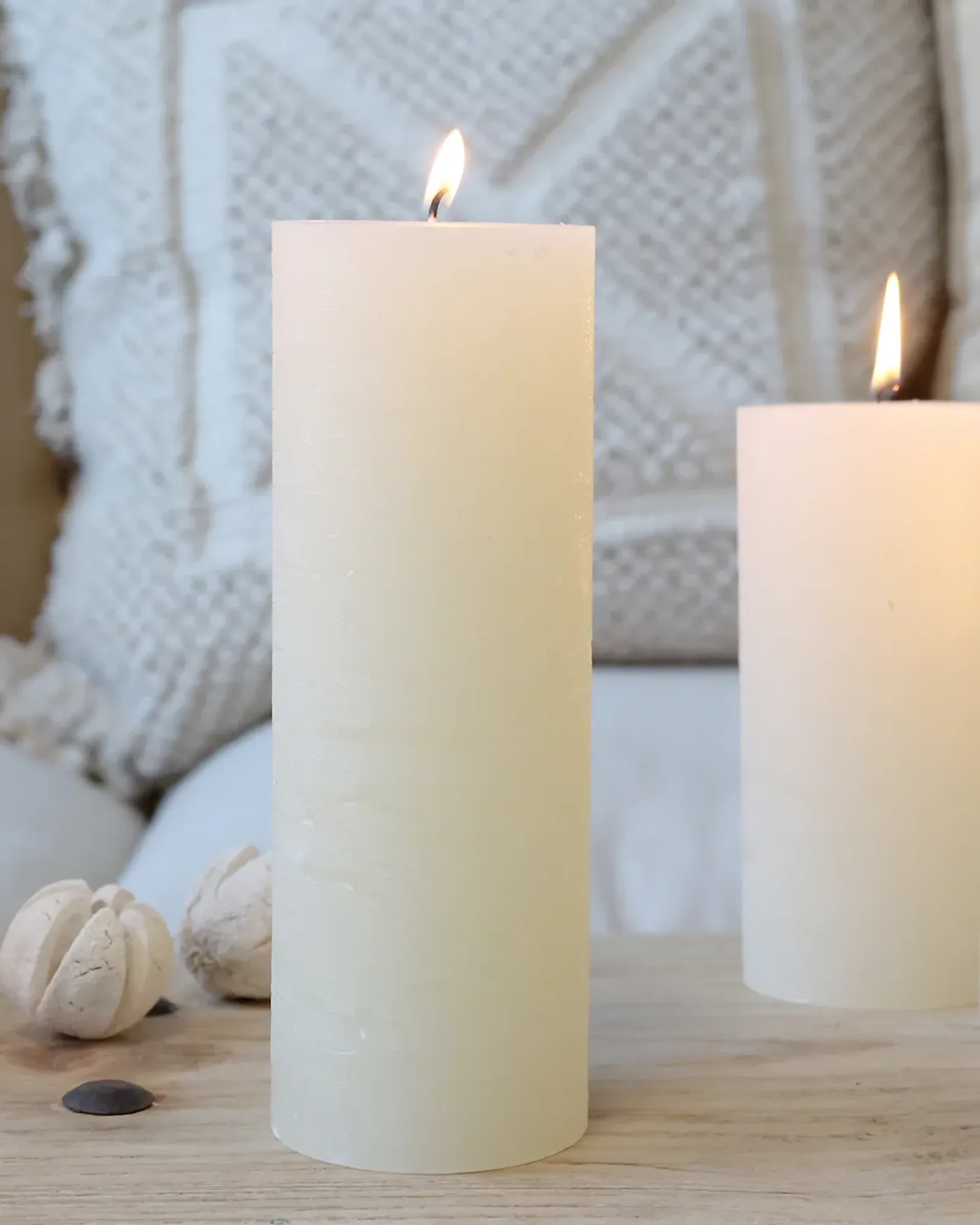 A tall, slim rustic pillar candle in cream, lit, with shorter candles of the same colour behind it.