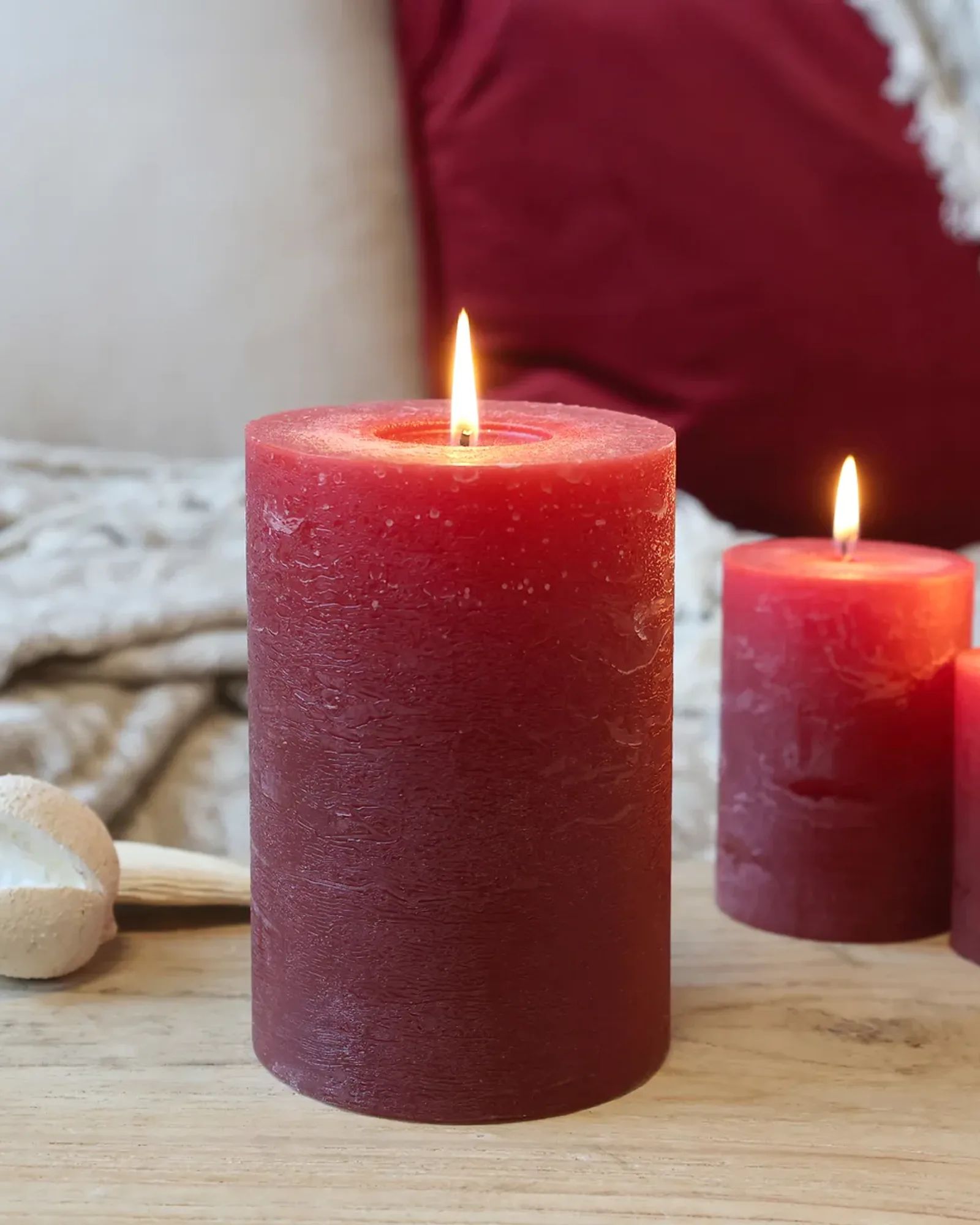 A short rustic pillar candle in dark red, lit, with taller candles of the same colour behind it.