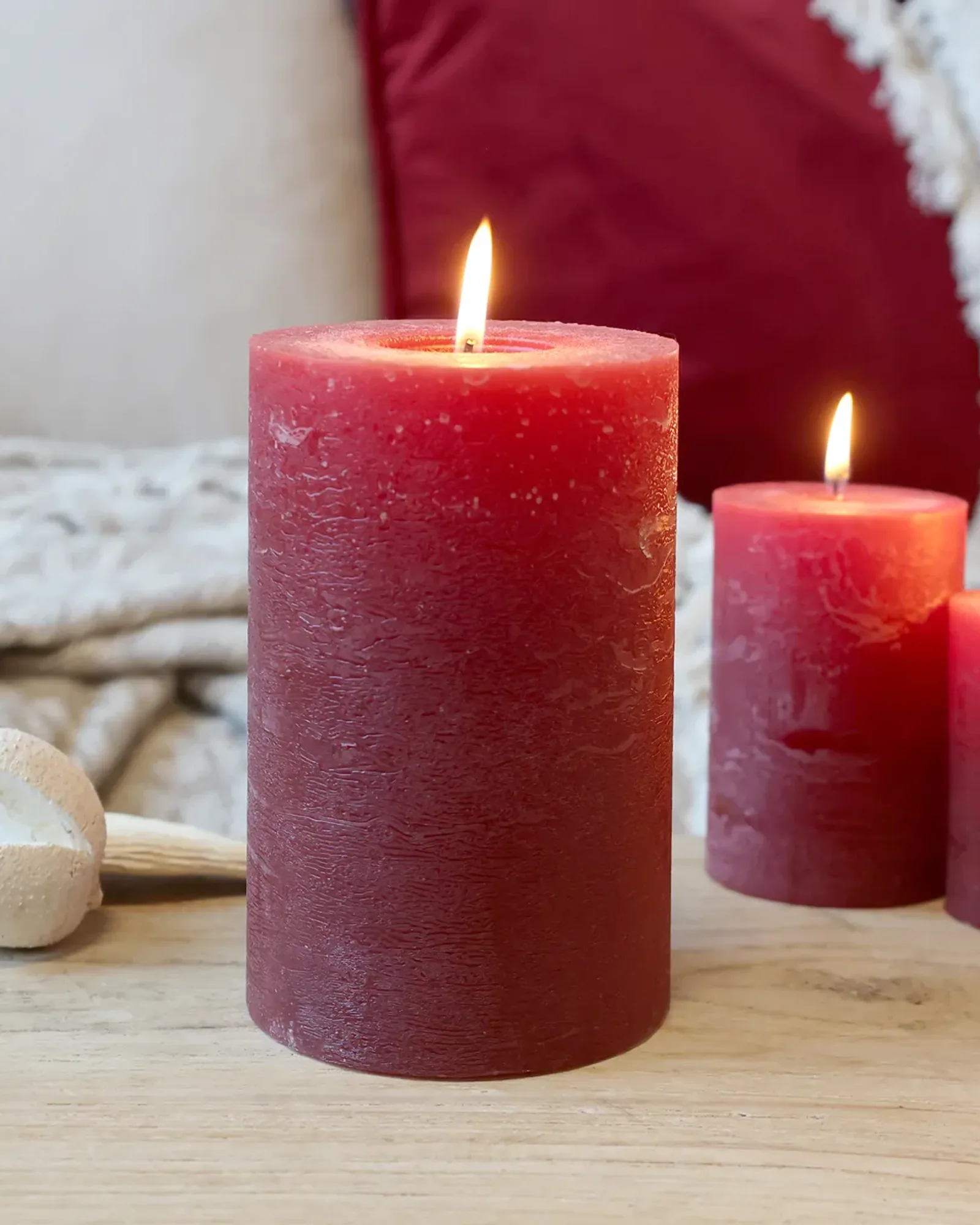 A wide rustic pillar candle in dark red, lit, with smaller candles of the same colour behind it.