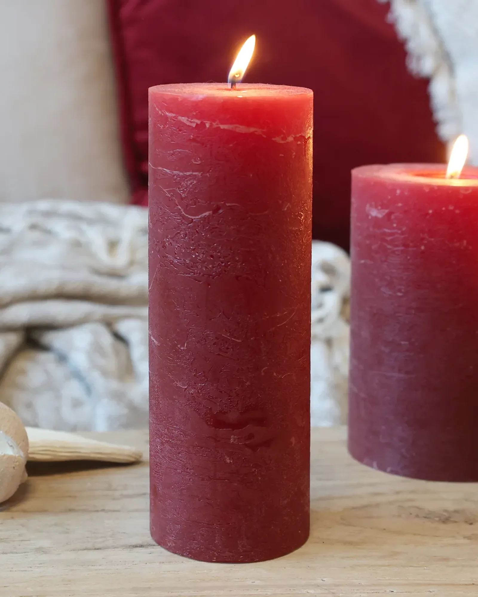 A tall, slim rustic pillar candle in dark red, lit, with shorter candles of the same colour behind it.