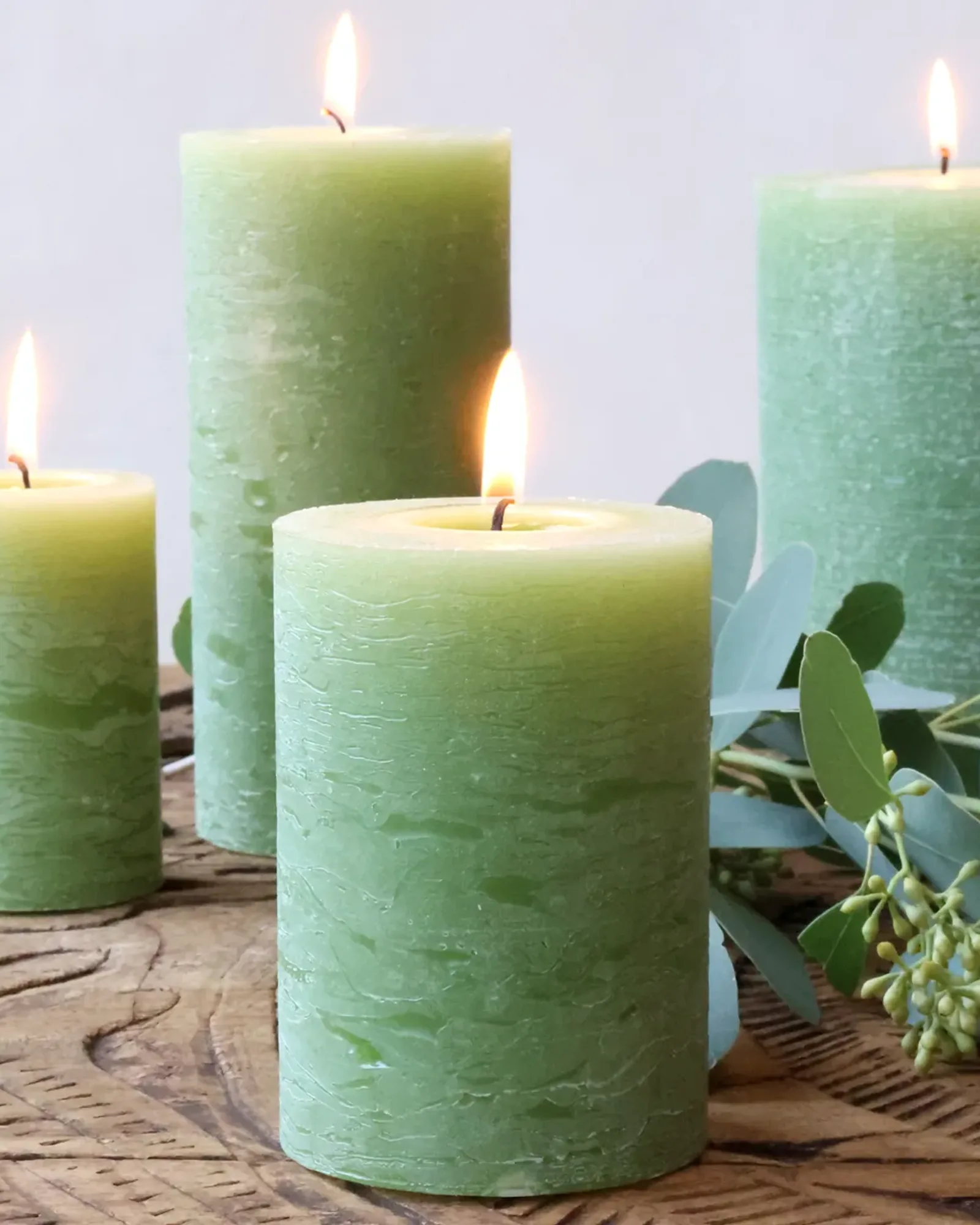 Four rustic pillar candles in a mid green in different sizes, with eucalyptus on a wooden board.