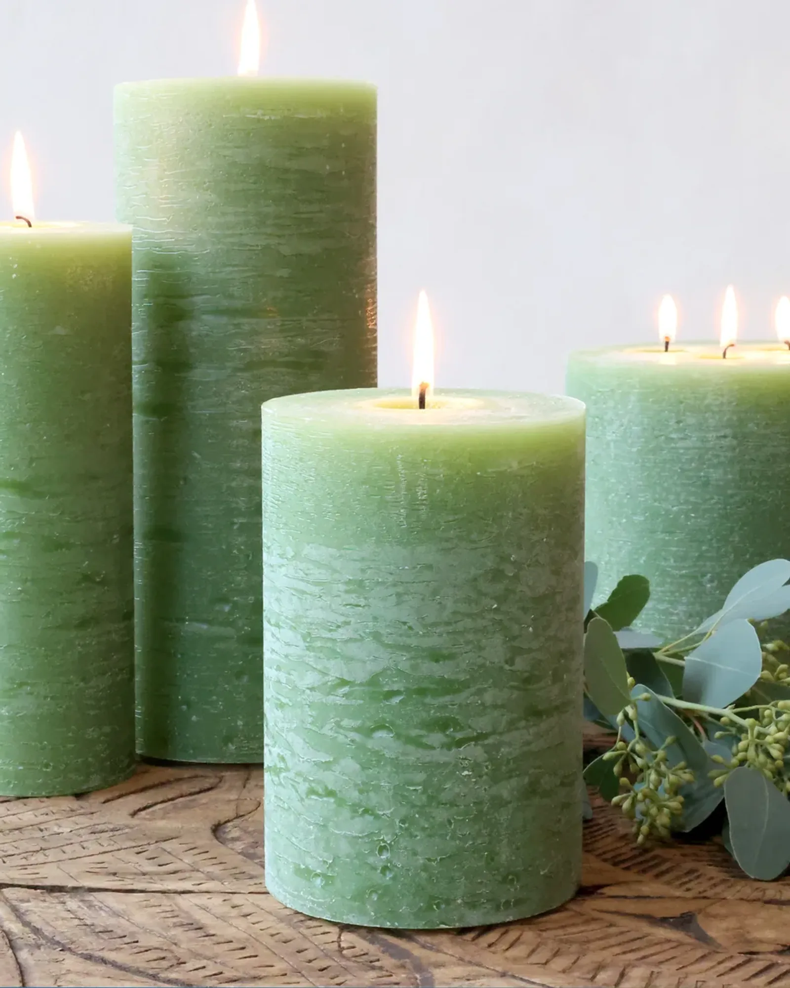 Four rustic pillar candles in a mid green in different sizes, with eucalyptus on a wooden board.