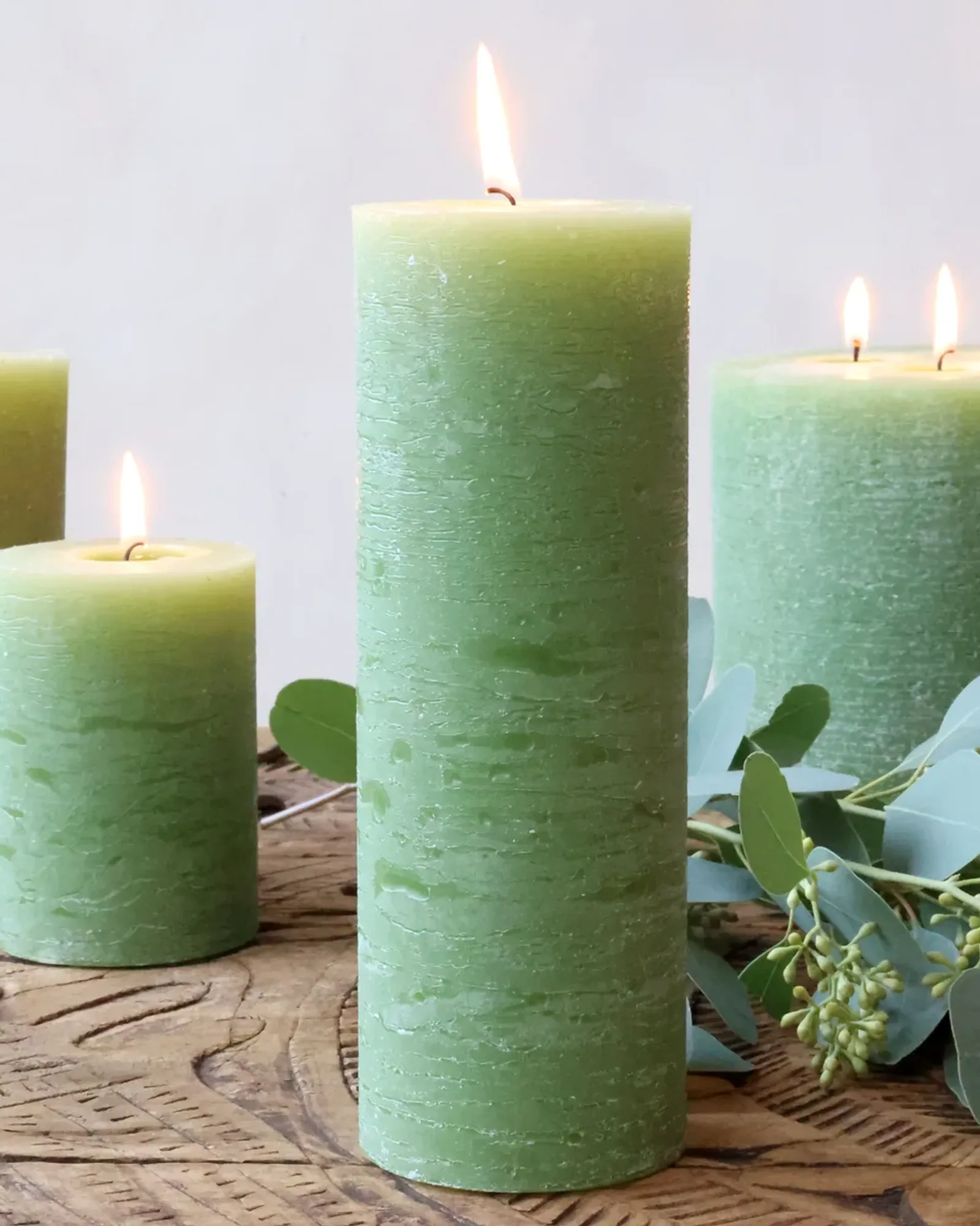 Four rustic pillar candles in a mid green in different sizes, with eucalyptus on a wooden board.