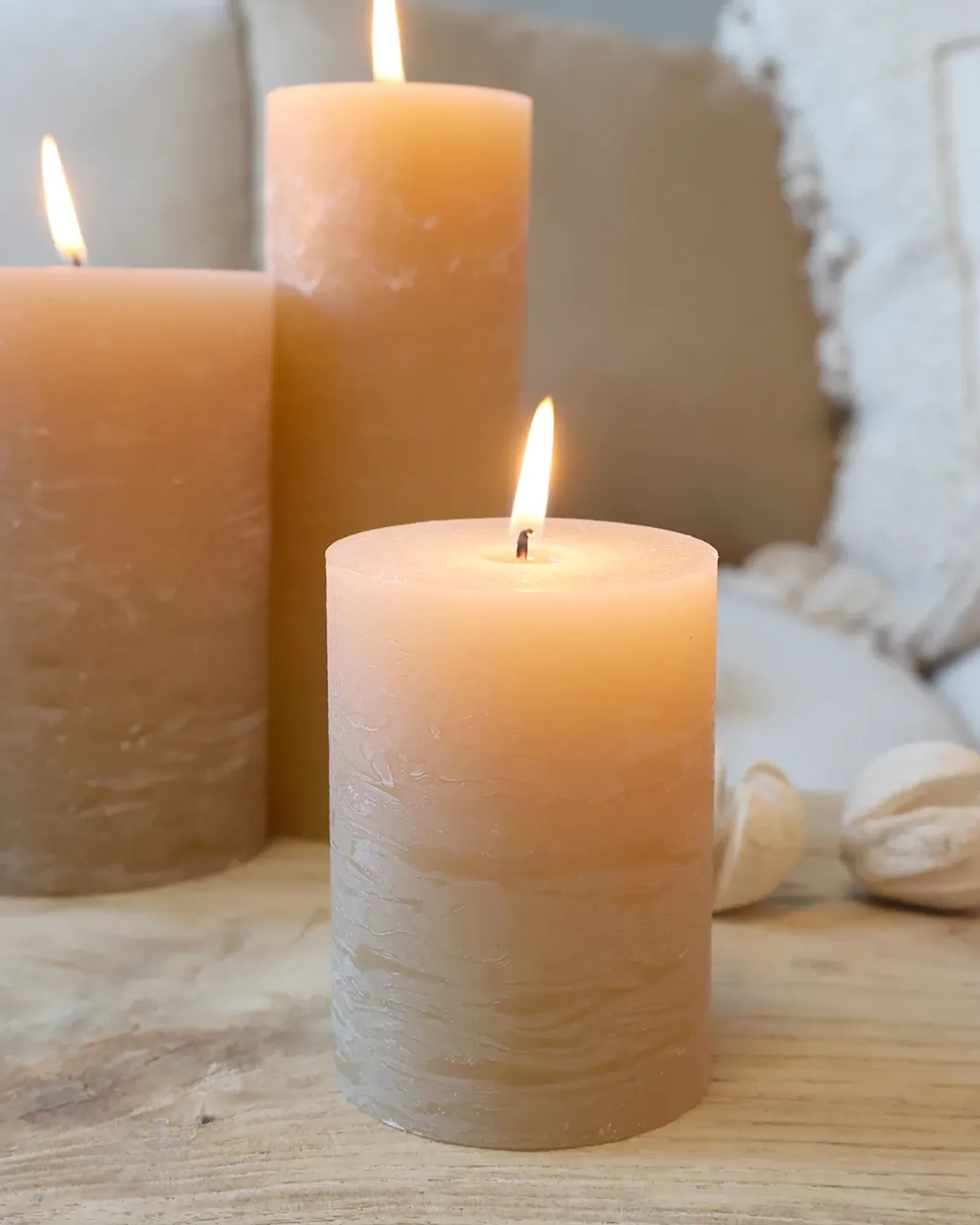 A short rustic pillar candle in honey, lit, with taller candles of the same colour behind it.
