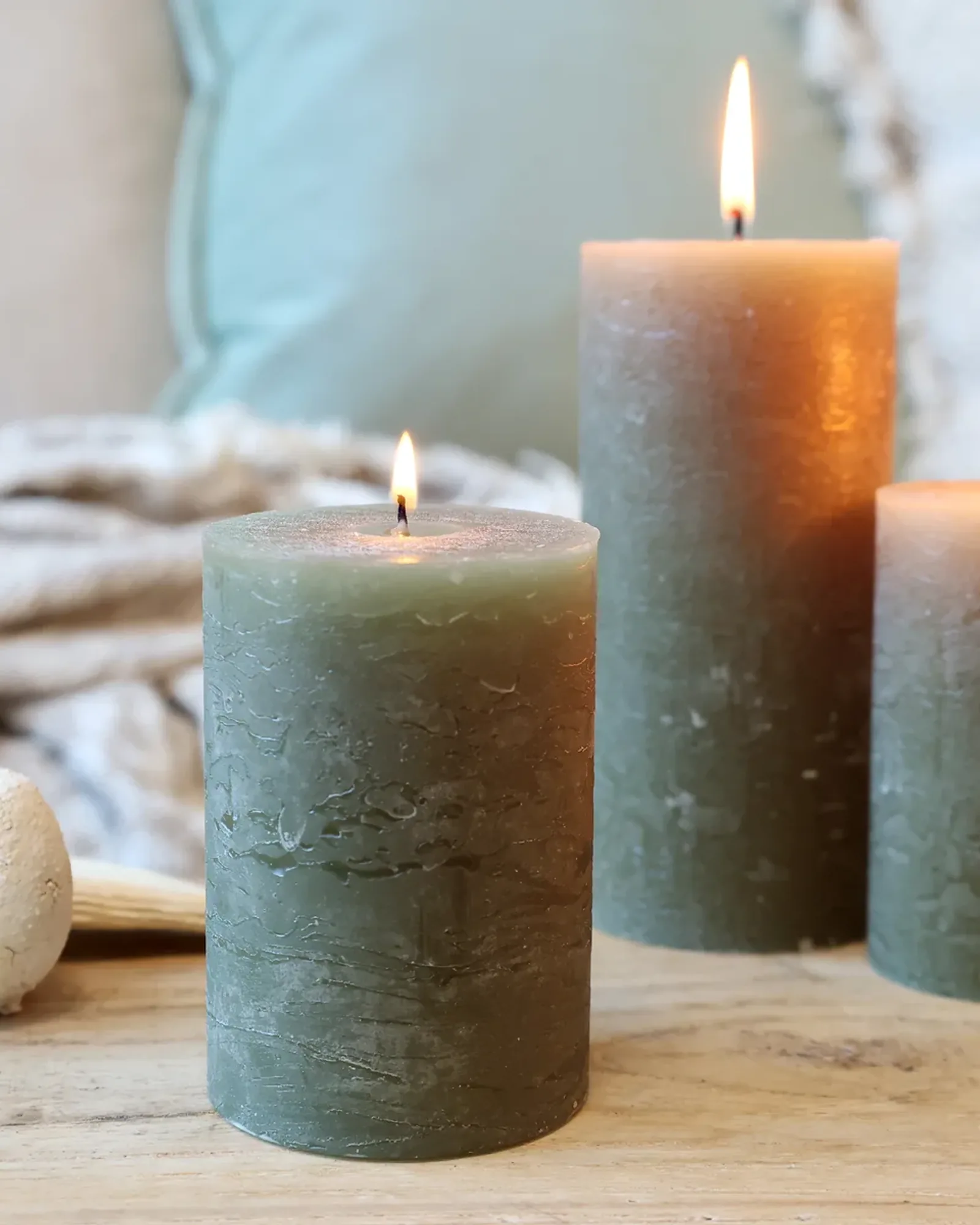 A short rustic pillar candle in olive green, lit, with taller candles of the same colour behind it.