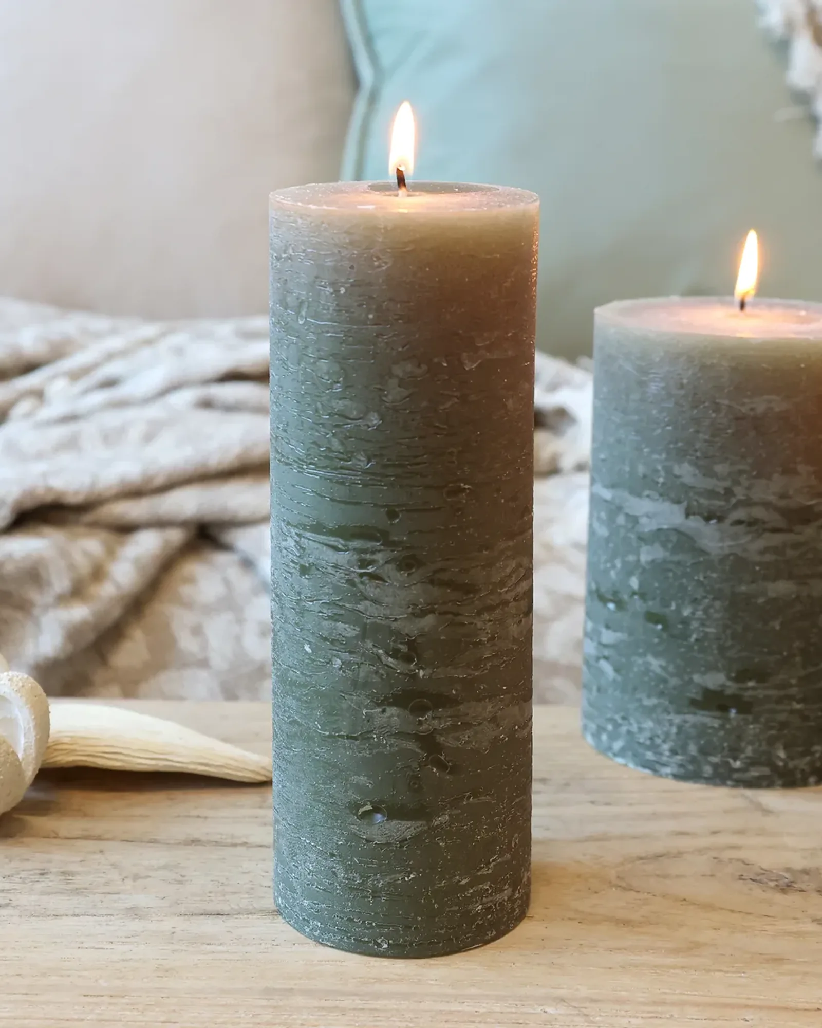 A tall, slim rustic pillar candle in olive green, lit, with shorter candles of the same colour behind it.