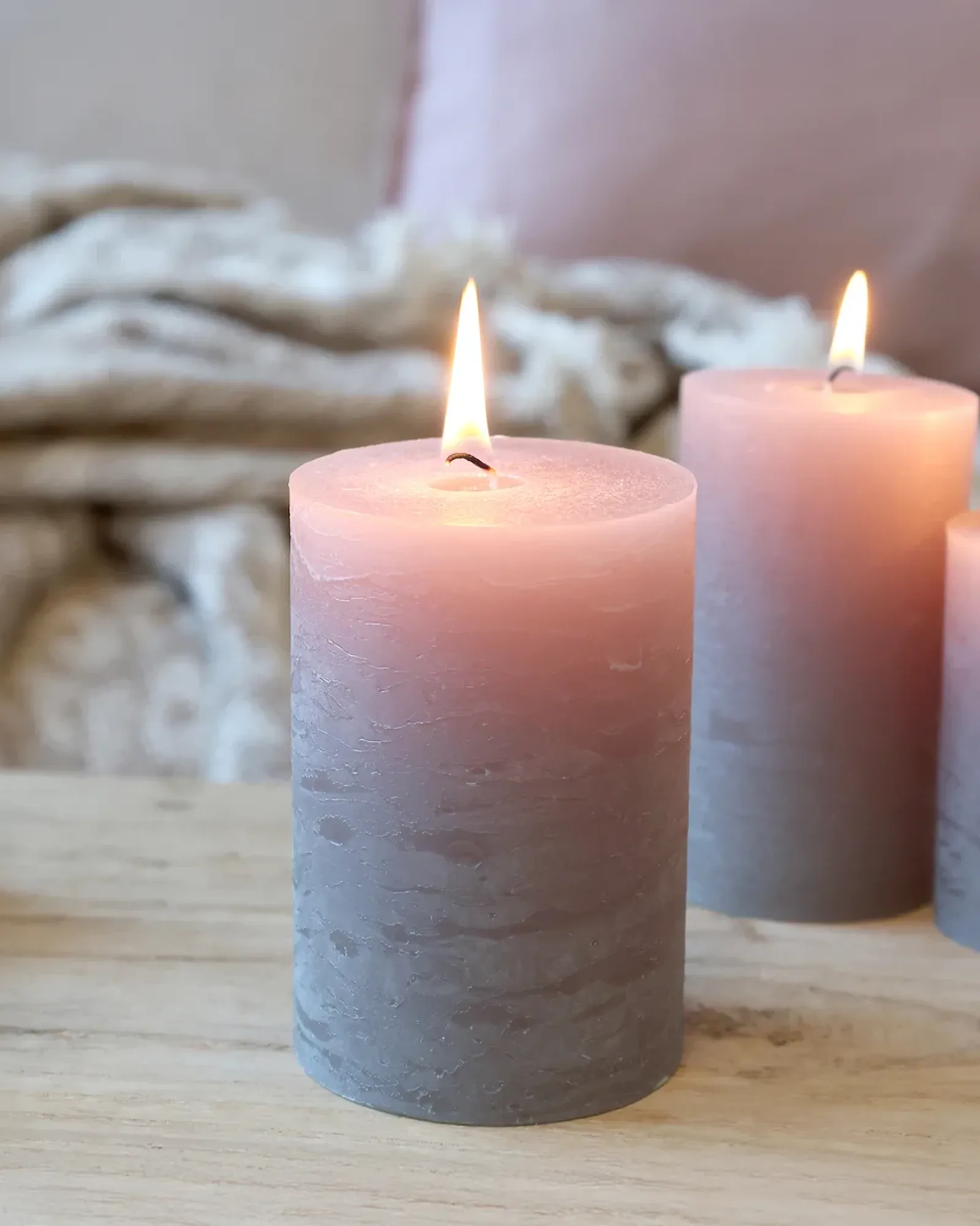 A short rustic pillar candle in taupe, lit, with taller candles of the same colour behind it.