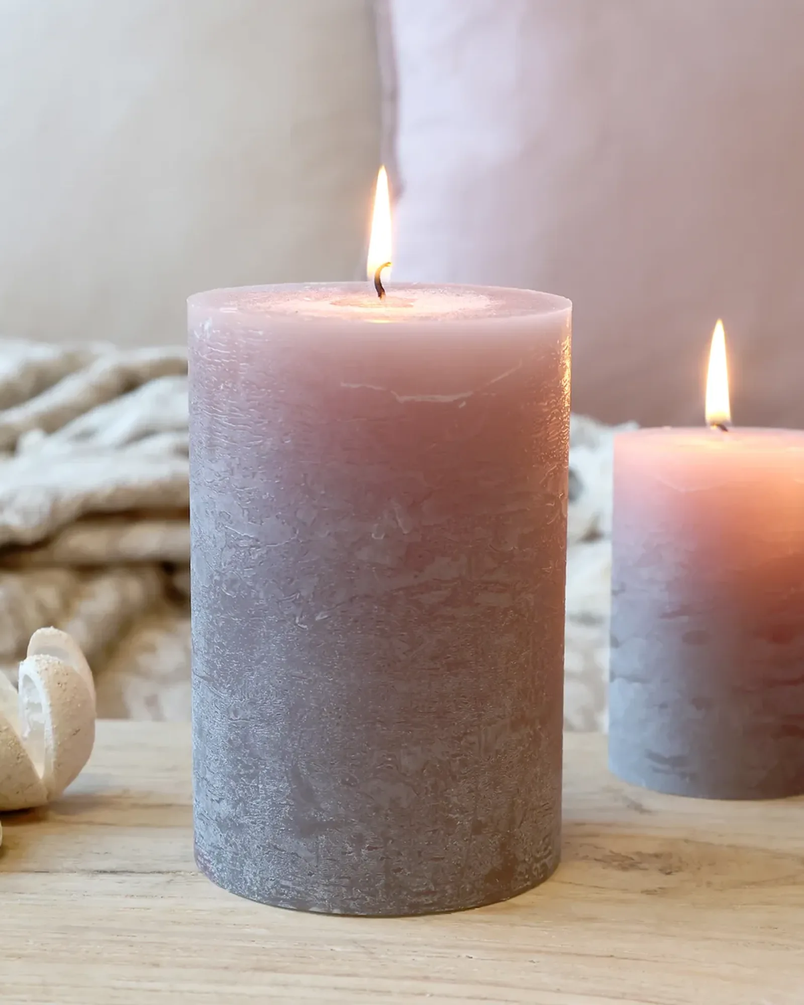 A wide rustic pillar candle in taupe, lit, with smaller candles of the same colour behind it.
