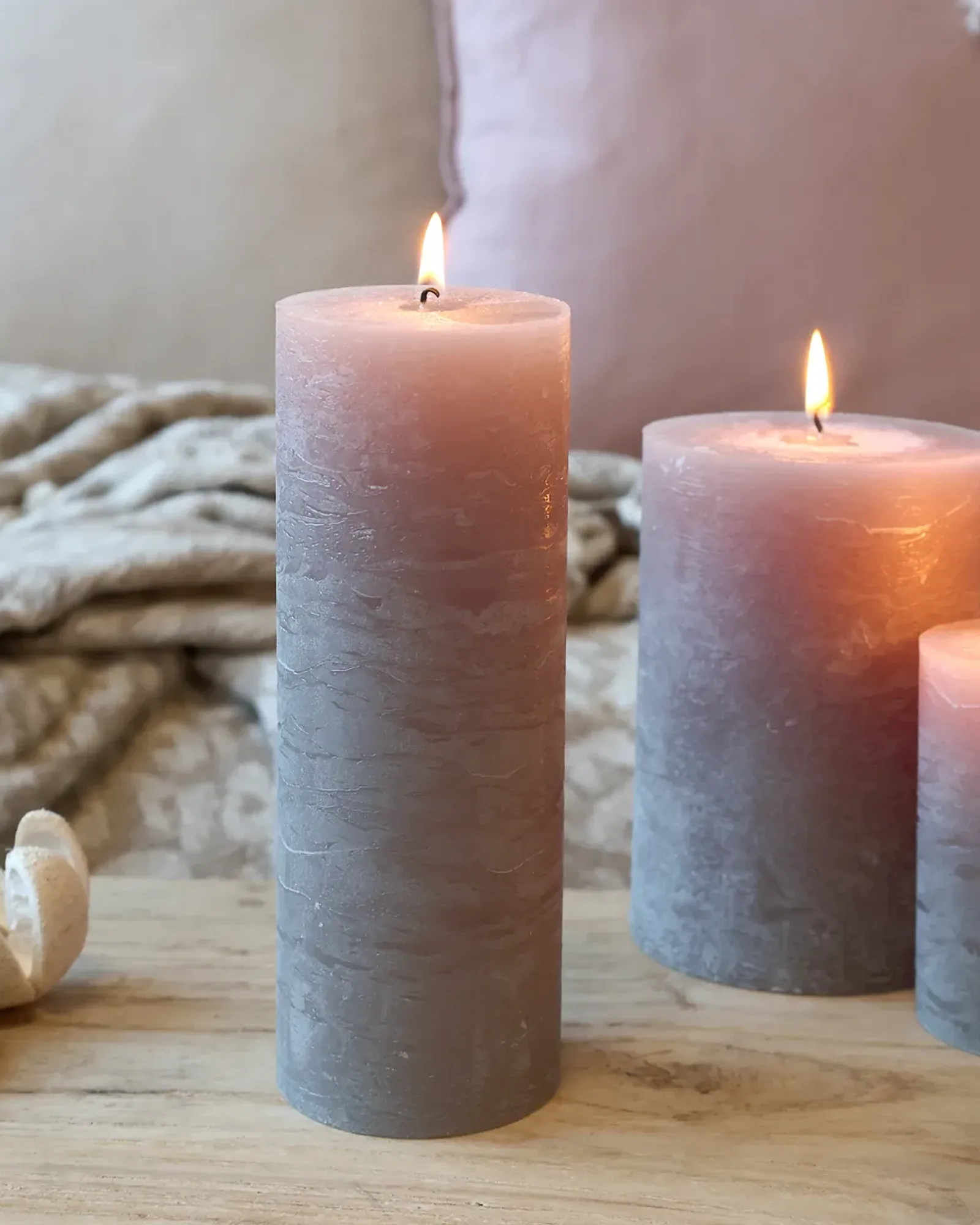 A tall, slim rustic pillar candle in taupe, lit, with shorter candles of the same colour behind it.