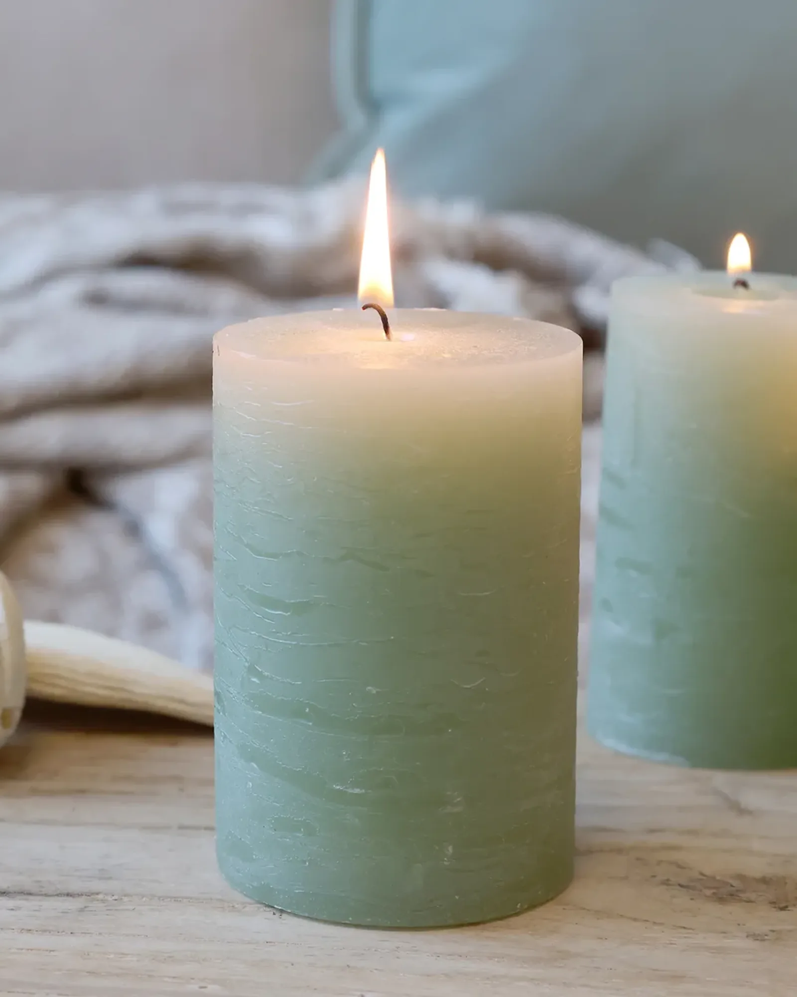 A short rustic pillar candle in sage green, lit, with taller candles of the same colour behind it.