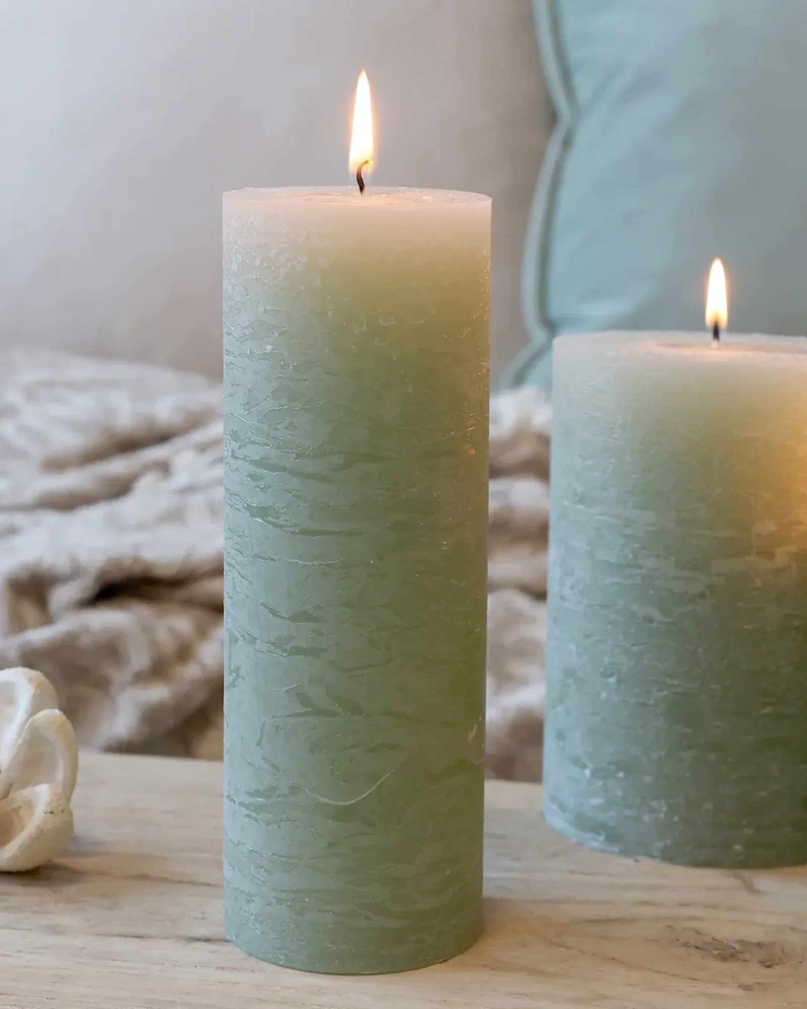 A tall, slim rustic pillar candle in sage green, lit, with shorter candles of the same colour behind it.