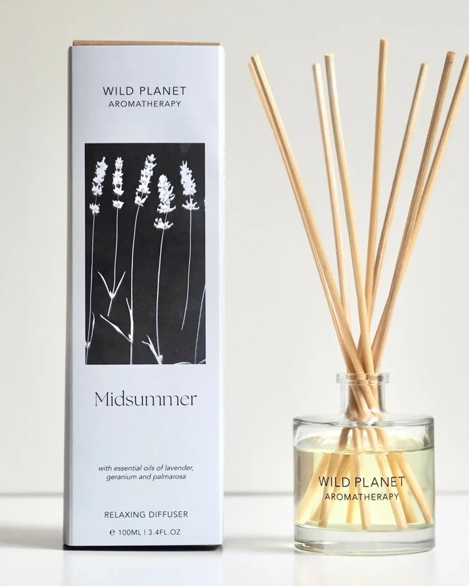 A clear glass bottle of Midsummer reed diffuser oil with a fan of natural reeds standing in it, beside its card box printed with the scent's name.