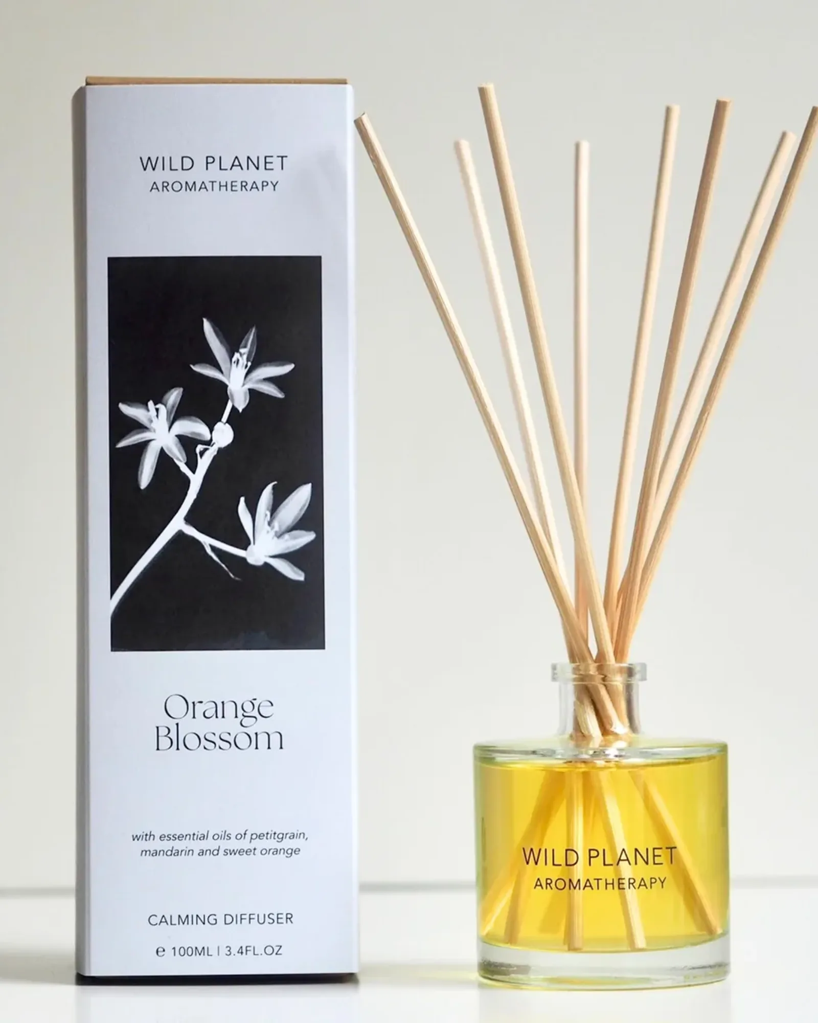 A clear glass bottle of Orange Blossom reed diffuser oil with a fan of natural reeds standing in it, beside its card box printed with the scent's name.