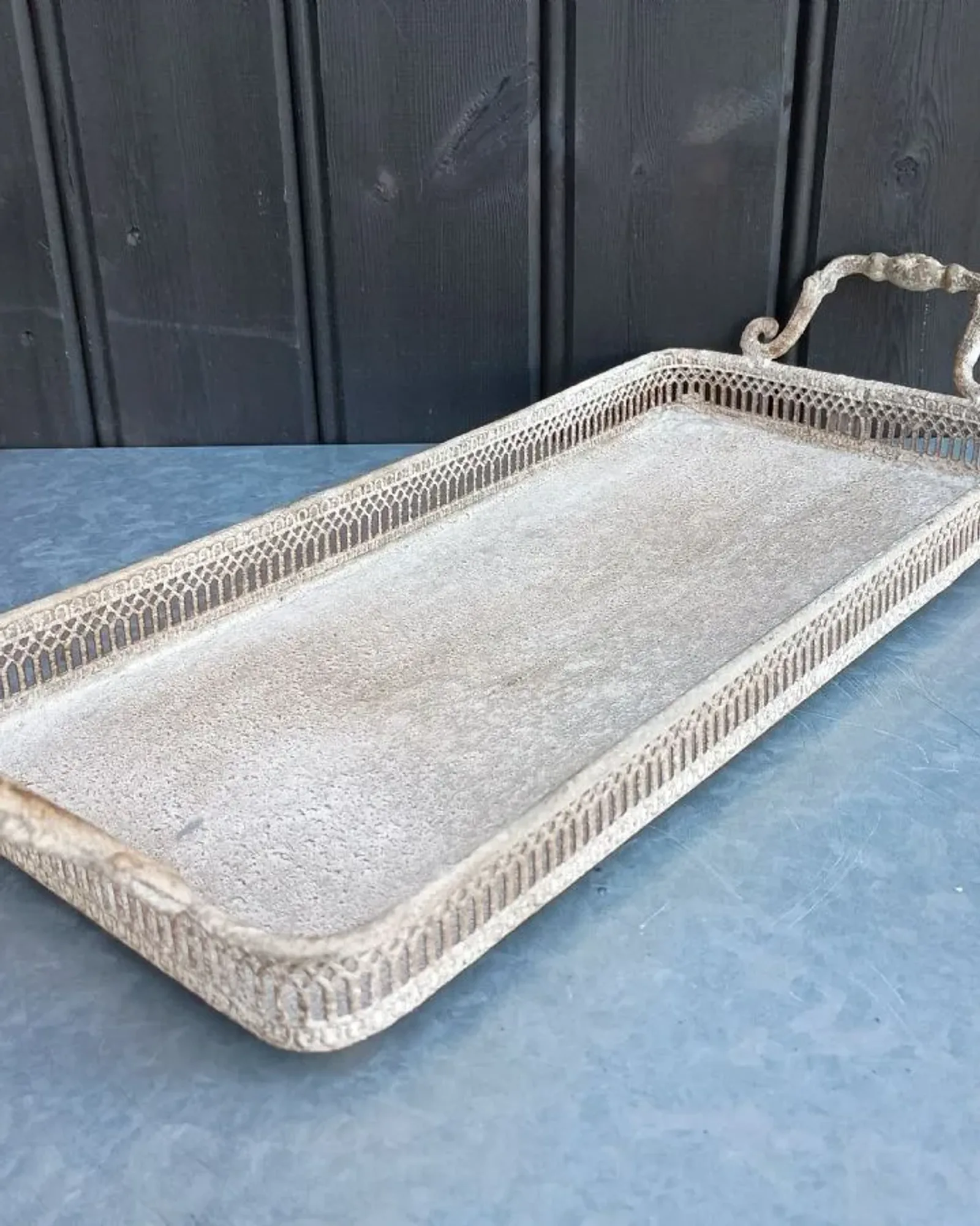 A long, narrow rectangular metal tray with a pierced gallery edge and two scrolled handles, in a distressed cream finish, on a blue table.