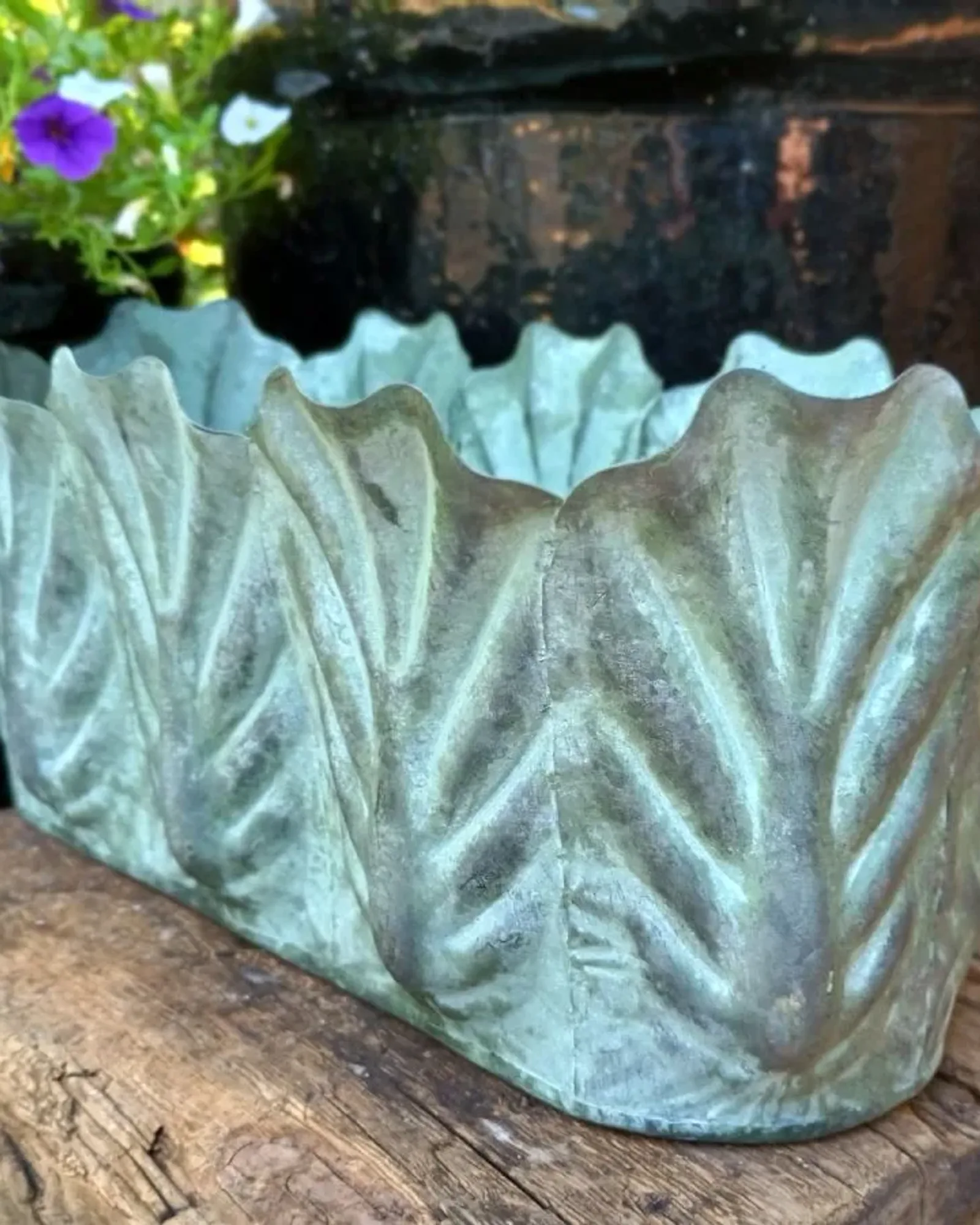 An oblong metal trough with scalloped, leaf-embossed sides, photographed in a garden with petunias and glazed pots behind.