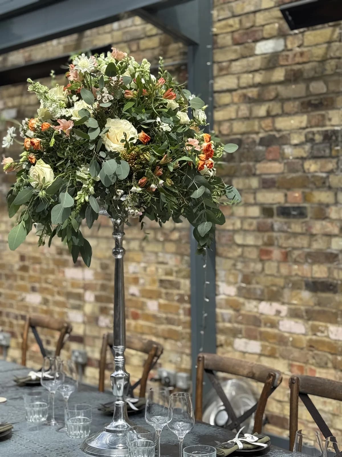 A tall pedestal arrangement of cream roses, orange spray roses and eucalyptus on a dining table against a brick wall
