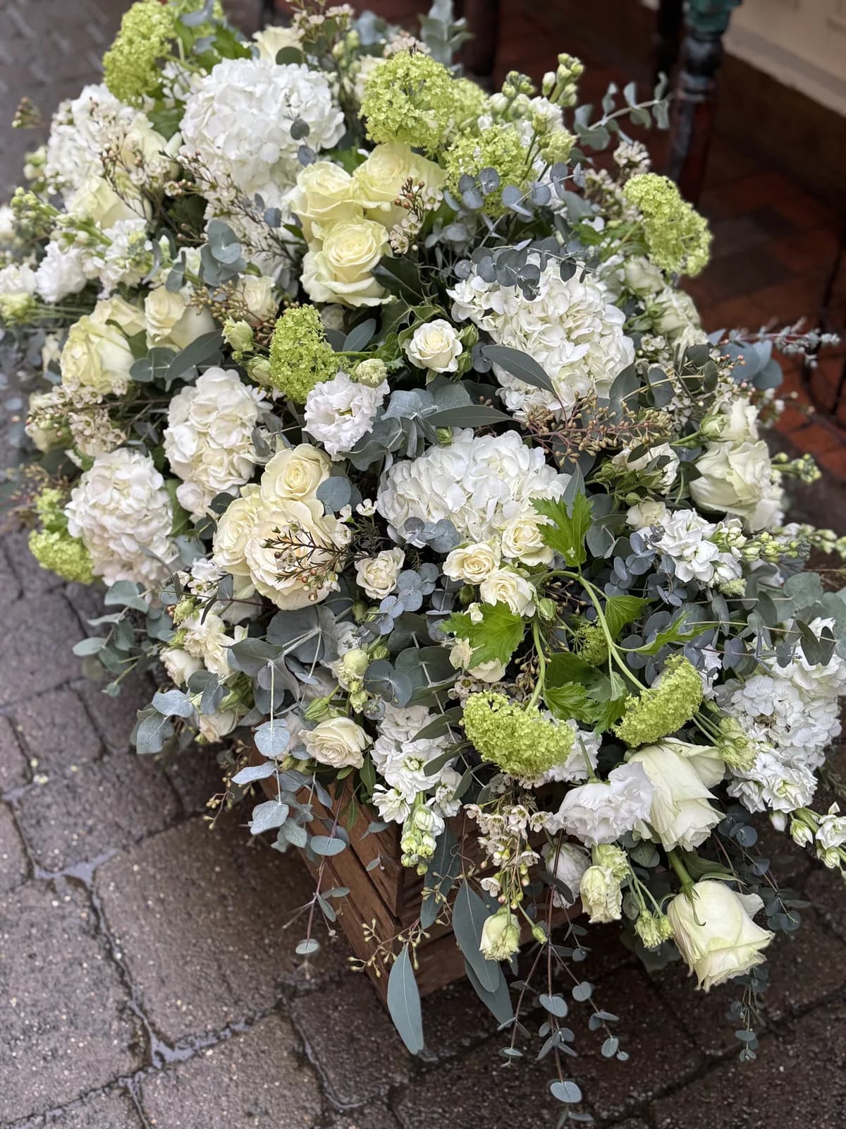 A large arrangement of white roses, hydrangea, stocks and lime-green guelder rose with trailing eucalyptus, set in a dark wooden crate