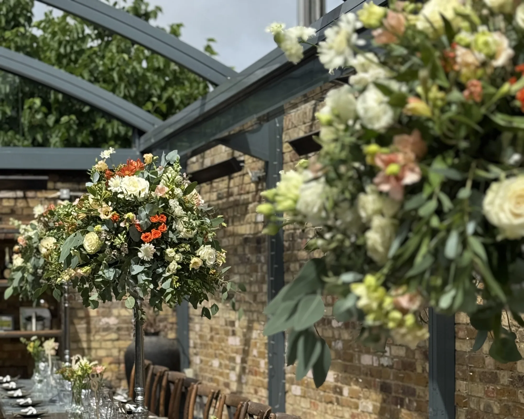 A long wedding table under a glass roof, dressed with tall Flower & Glory arrangements and bud vases